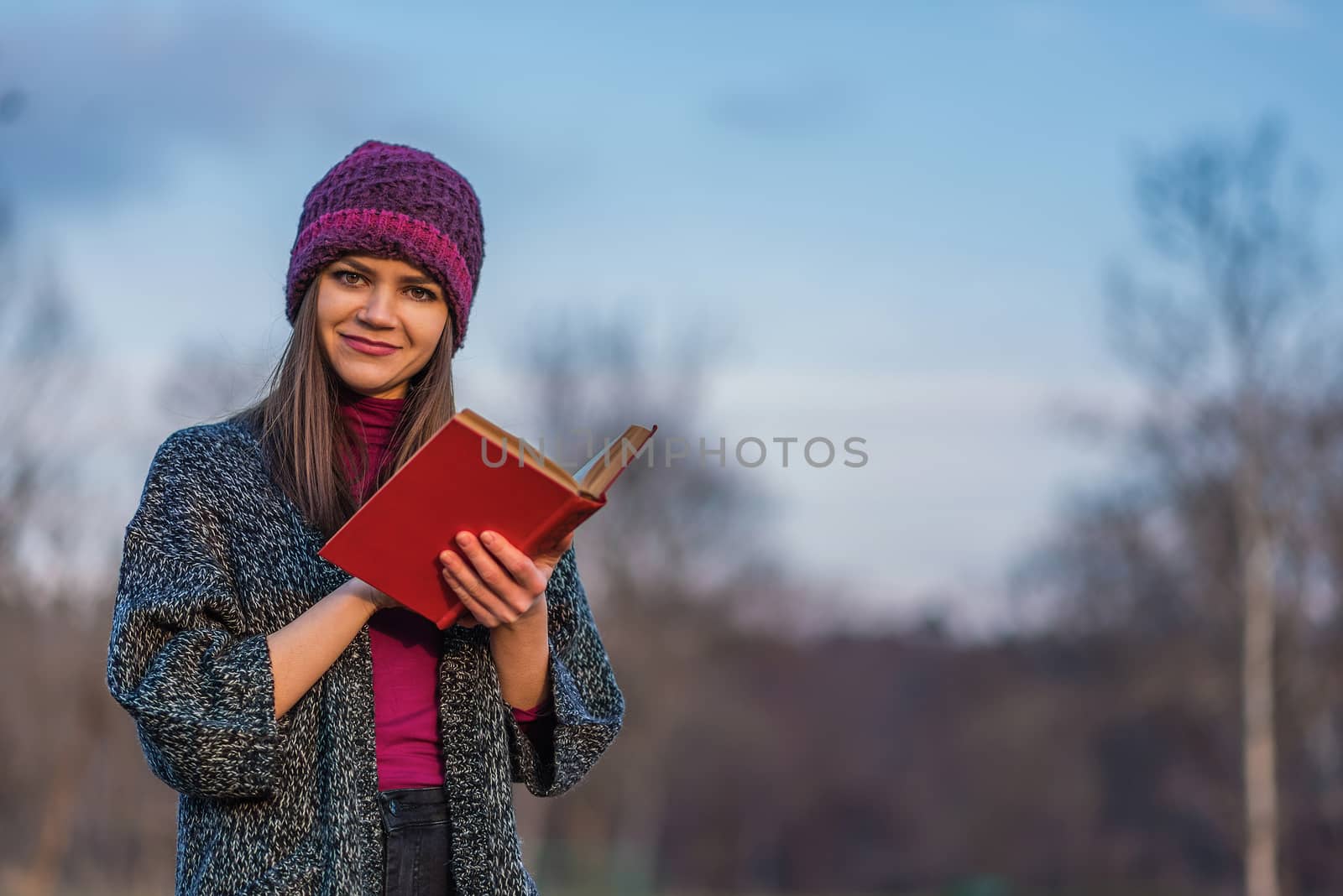A teenager girl in purple hat holding opened book with red cover while standing in a park.