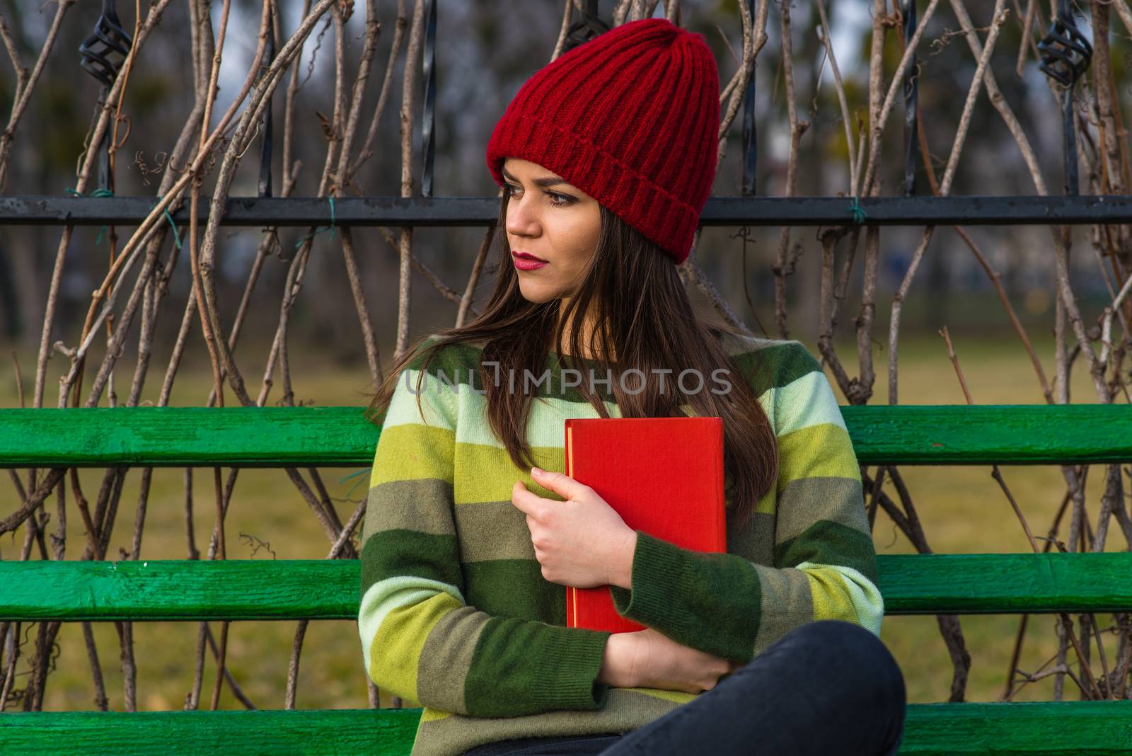 Teenager girl in red hat and green sweater sitting on a bench in a park and holding a red book.