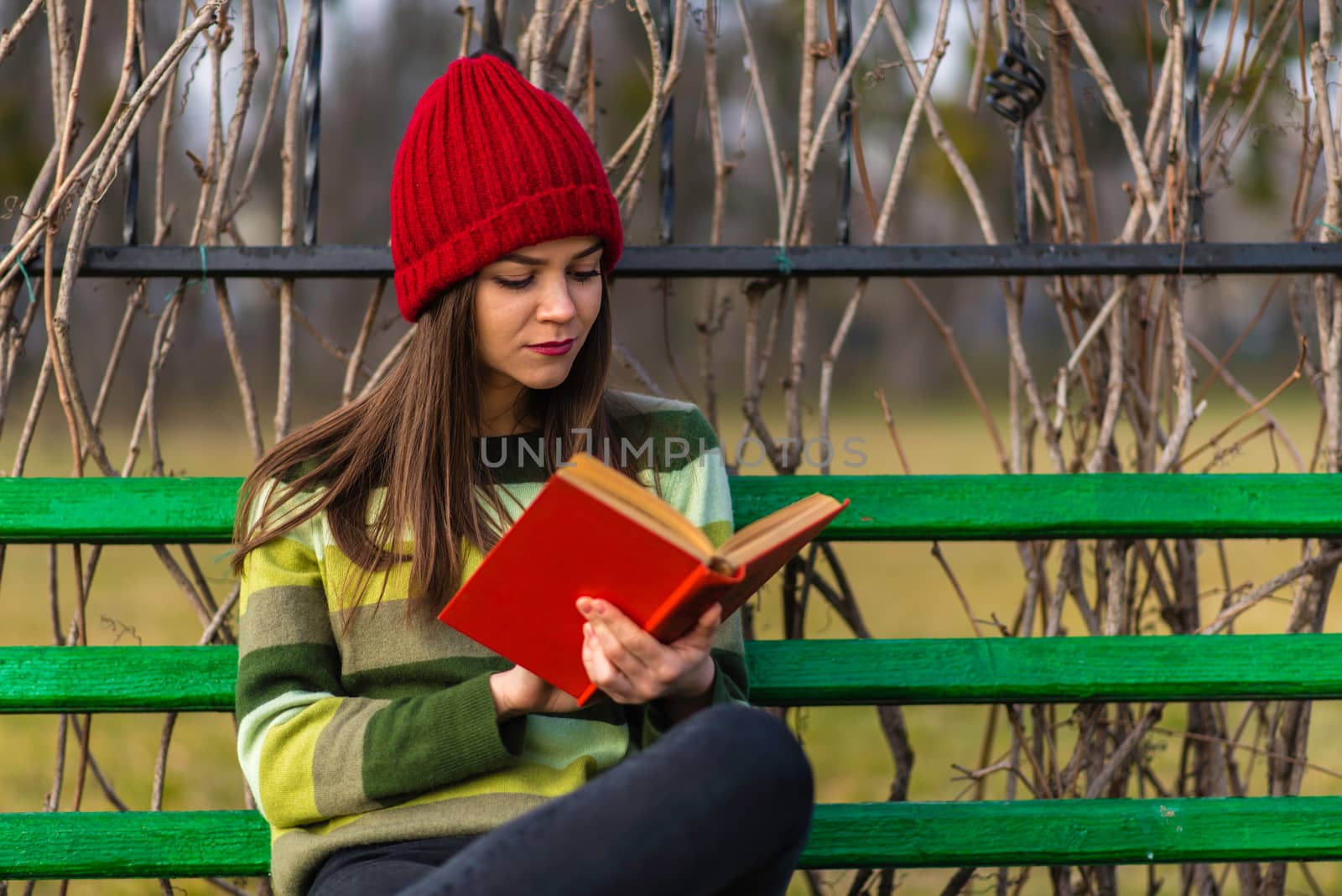 Teenager girl in red hat and green sweater sitting on a bench in a park and reading a book with red cover.