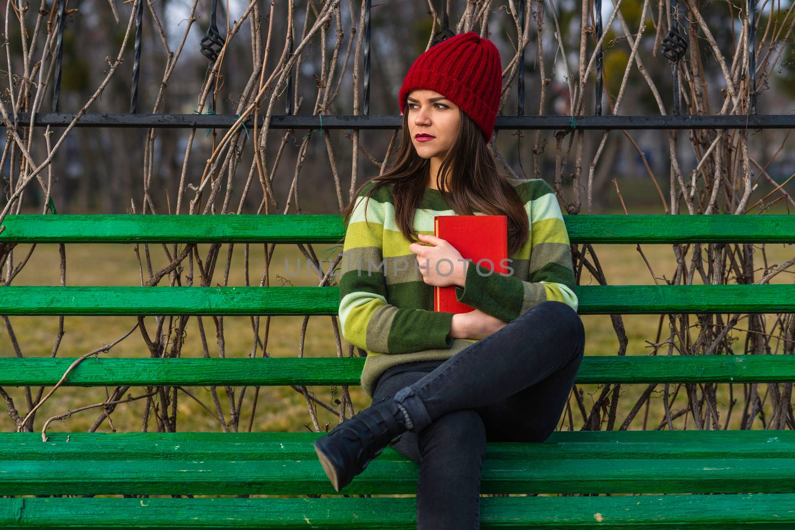 Teenager girl in red hat and green sweater sitting on a bench in a park and holding a red book. Medium shot.