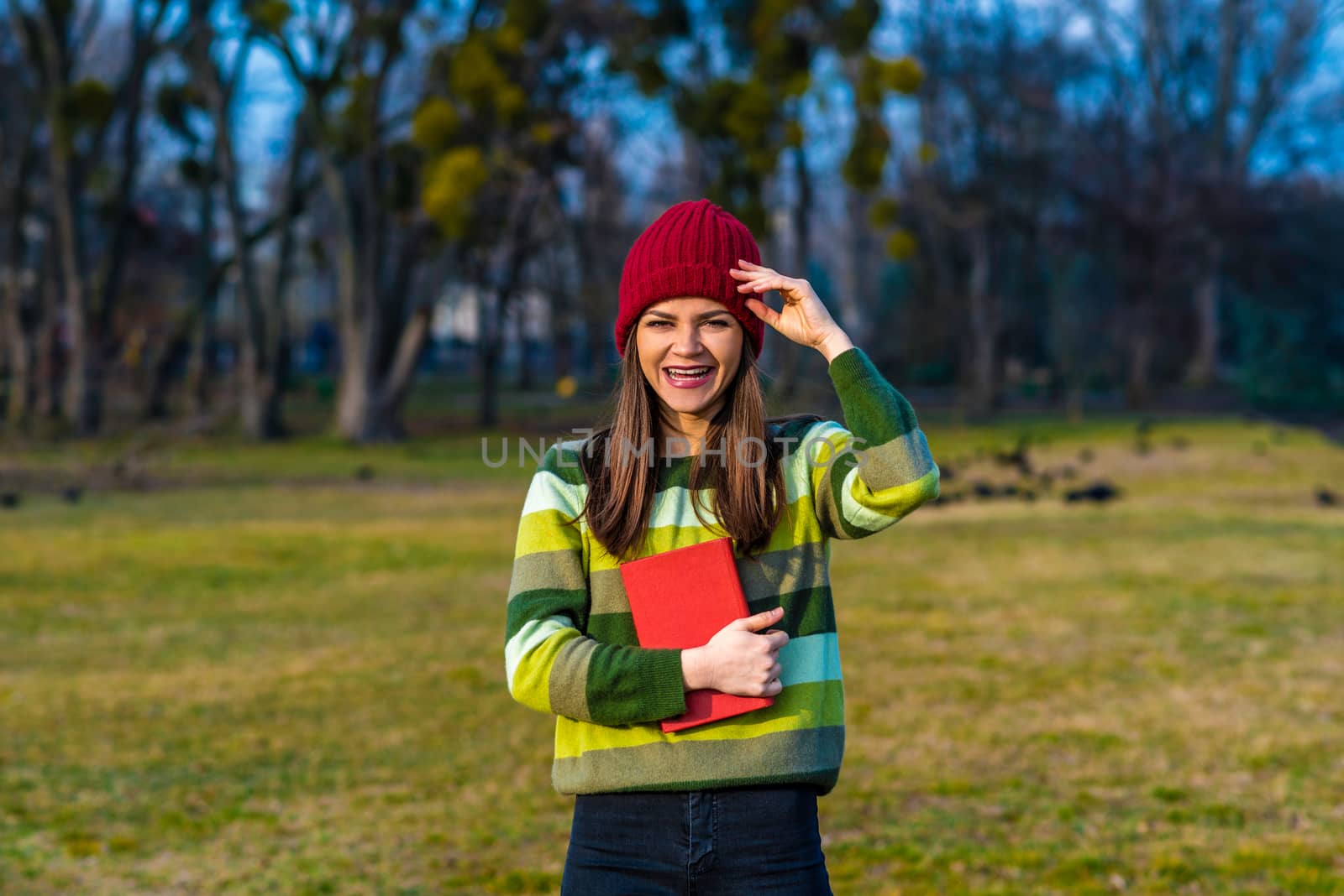 A teenager girl in red hat and green striped sweater is laughing andholding book with red cover while standing on a lawn in a park