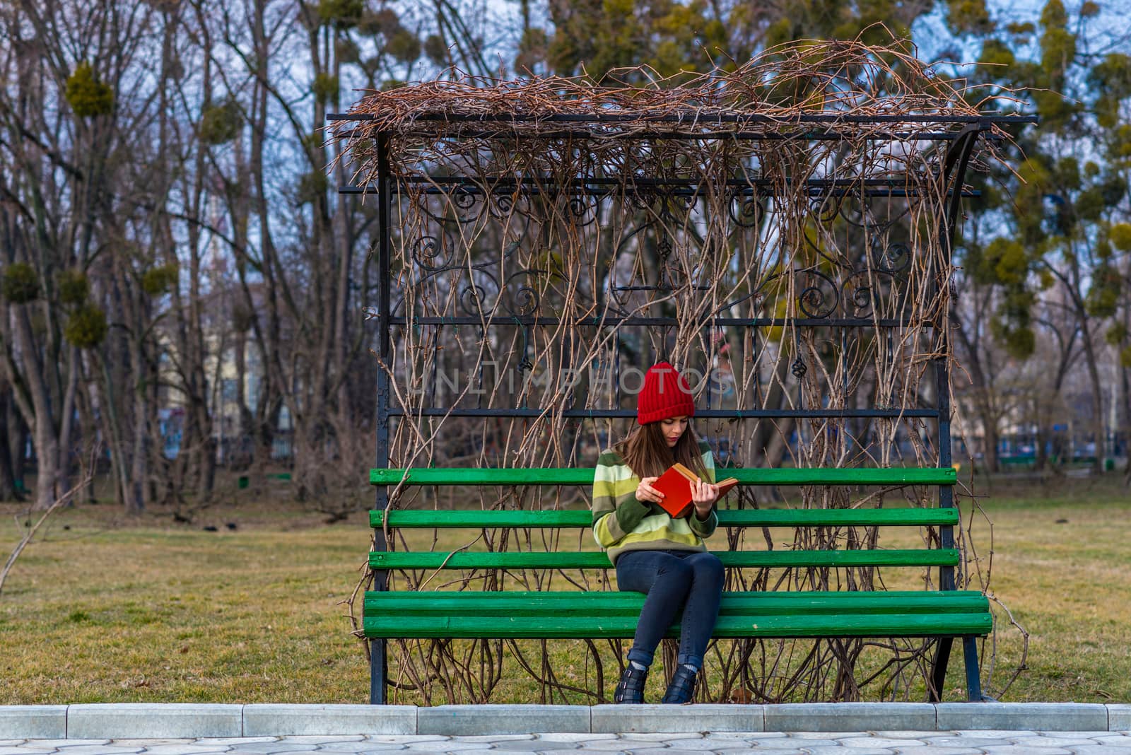 A teenager girl is sitting on a bench in a park and reading book with red cover.