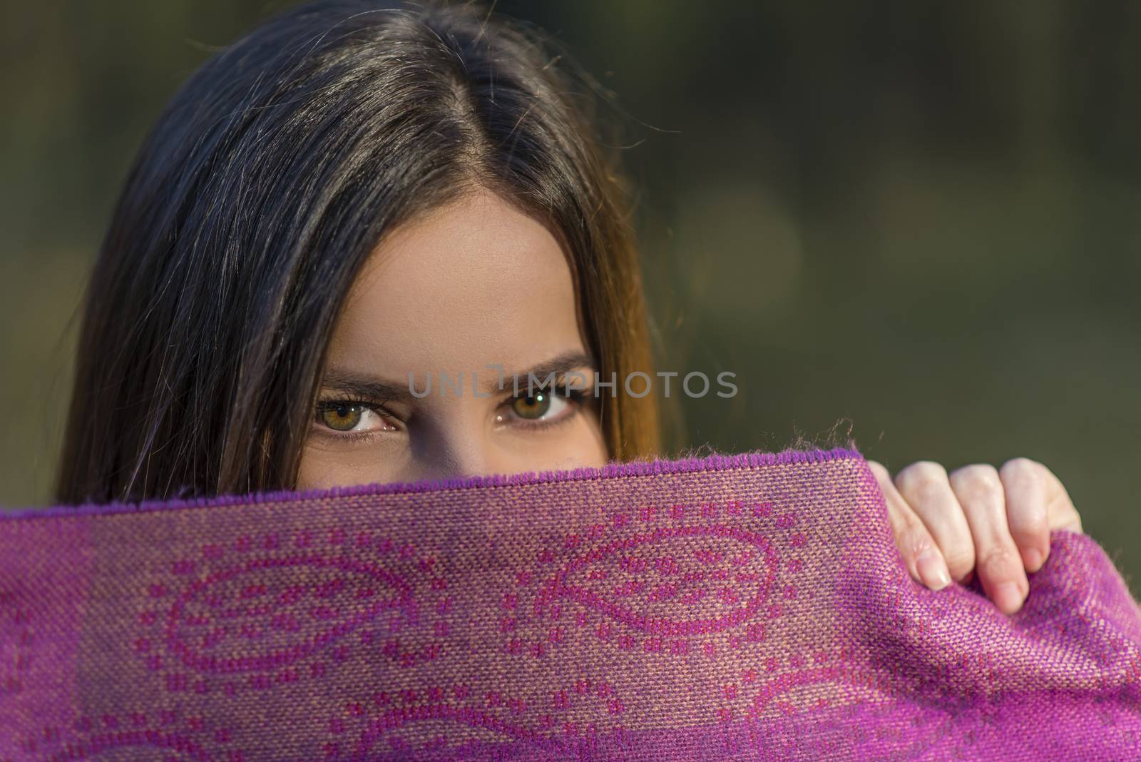 Eyes above the purple scarf. Pretty teenager girl with brown eyes is covering her face with the purple scarf in the afternoon woods. Shallow depth of field.