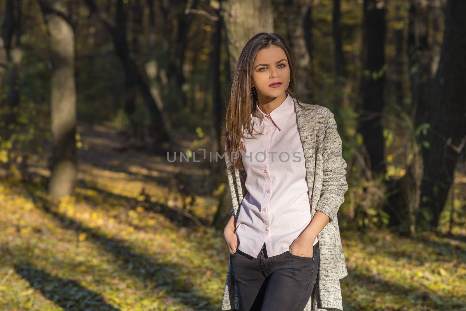A pretty teenager girl is posing in an autumn oak forest at afternoon. She has a detached and thoughtful expression on her face. She is holding her hands in her jeans pockets. 