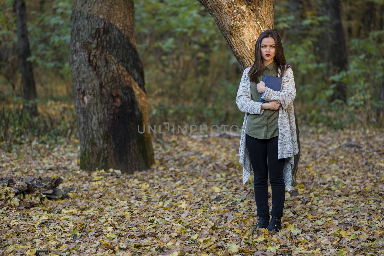 Scared by reading fairytales. A teenager girl is holding a book in an evening forest with two oak trees in the background. She is standing on foliage. The girl has a serious expression on her face.