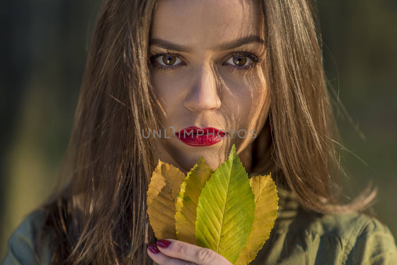 A close-up portrait of a teenager girl holding autumn yellow leaves in her hands. Gir has red lips, brown eyes and cold look.