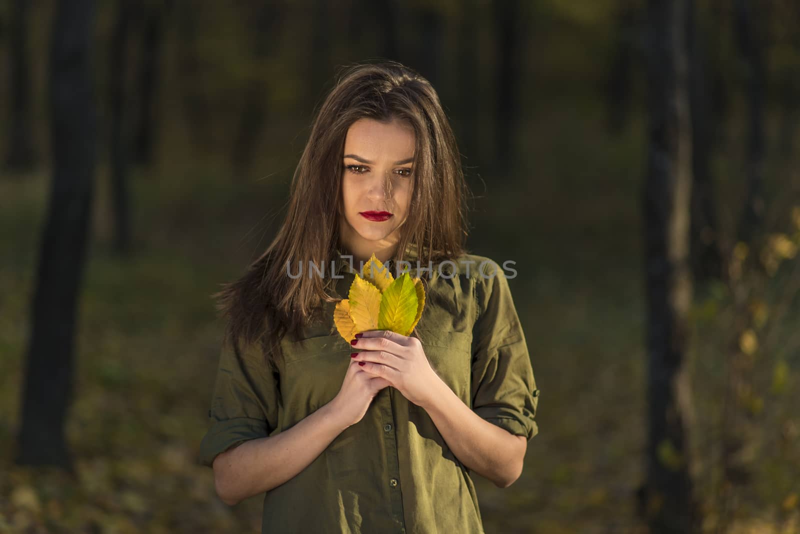 Pensive autumn look. A meditative teenage girl is looking down in a autumn forest while holding yellow leaves in her hands.
