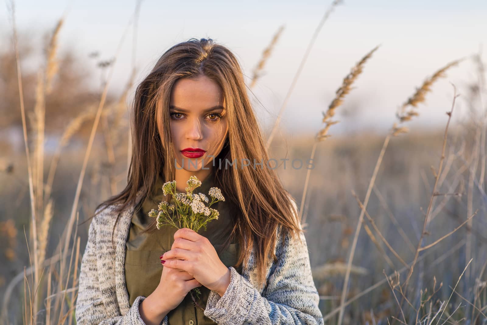 A beautiful teenage girl sitting sad in a field with a bouquet of field flowers in hands at autumn sunset. Girl has brown eyes and hair and red lips. 