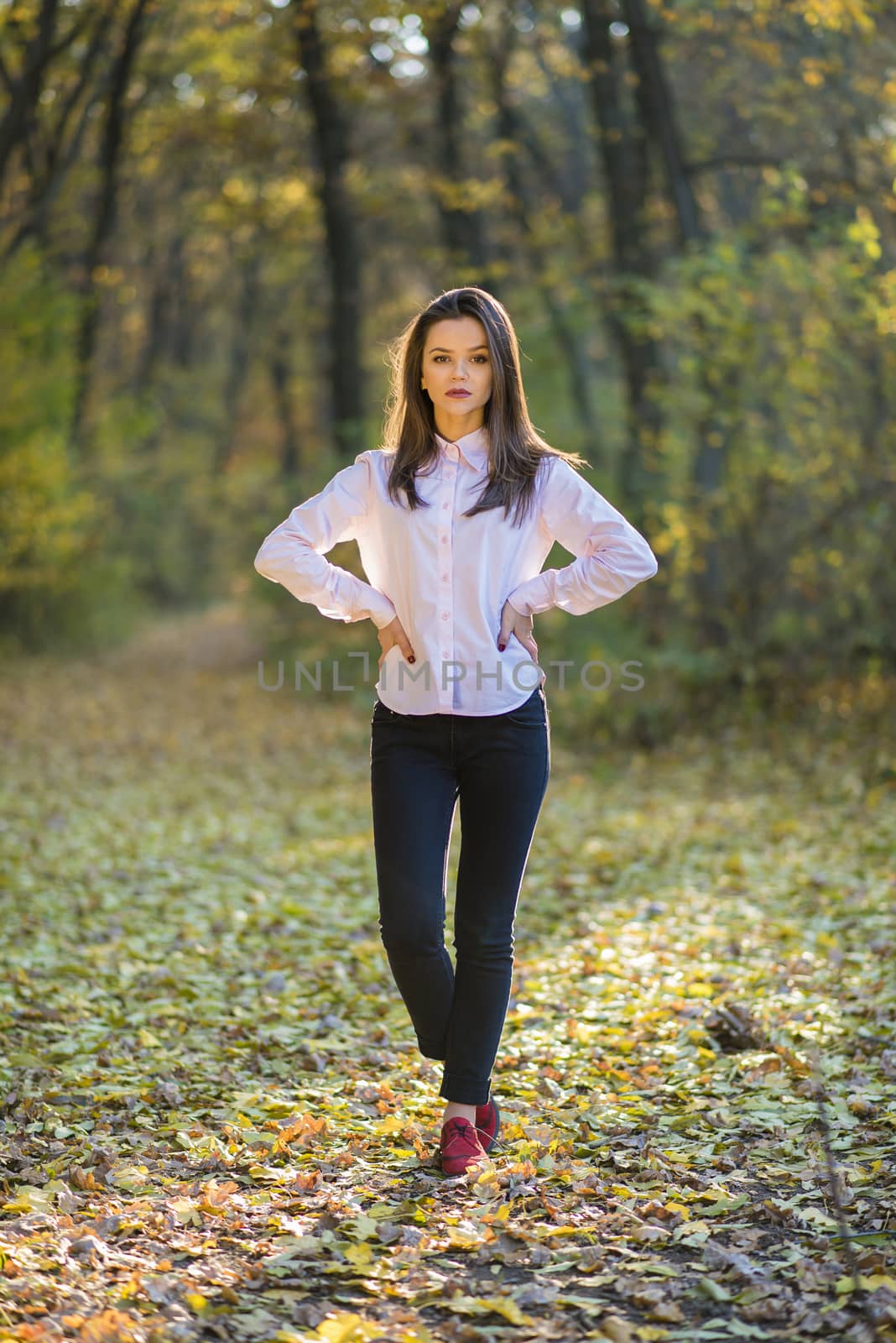 Posing on footpath in forest. A serious looking girls is posing in a pink shirt, dark blue jeans and red shoes in an autumn forest while standing on foliage.