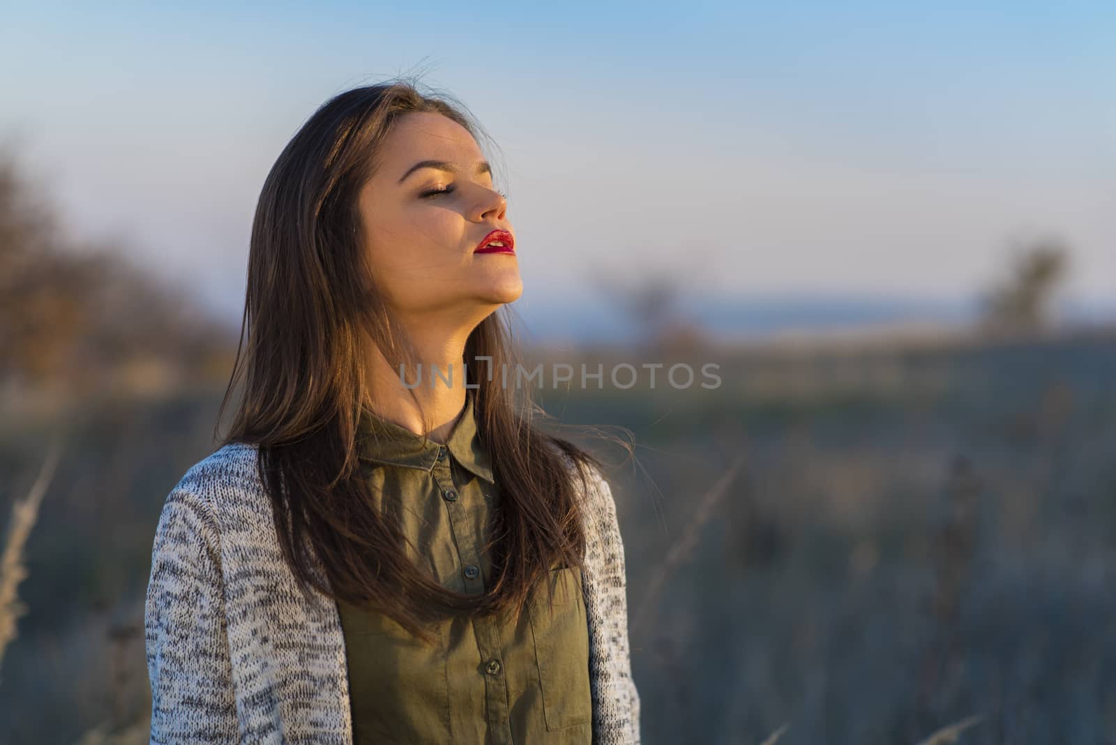 Enjoying sun before winter. A beautiful teenage girl standing in a field at autumn sunset, enjoying last sun days before winter. Girl has brown hair and red lips.