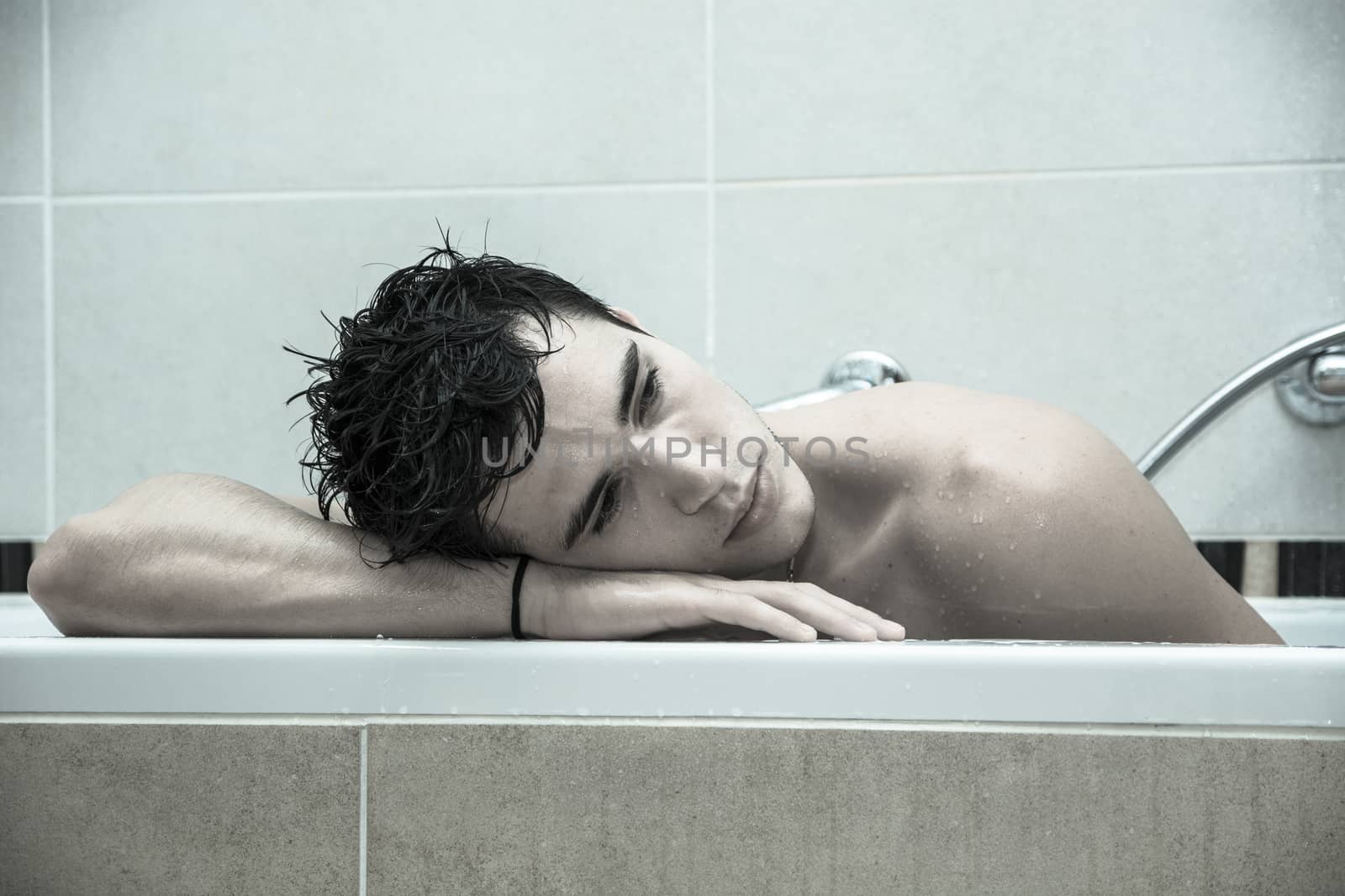 Handsome young man in bathtub at home having bath, washing body and hair with bathfoam and shampoo