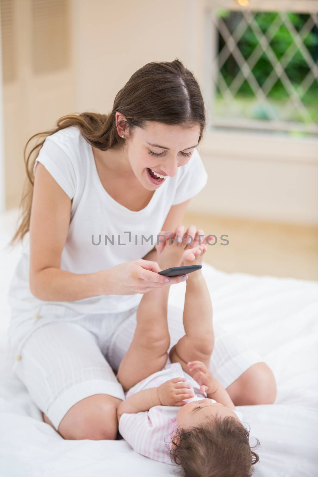 Smiling mother using mobile phone while playing with baby on bed at home