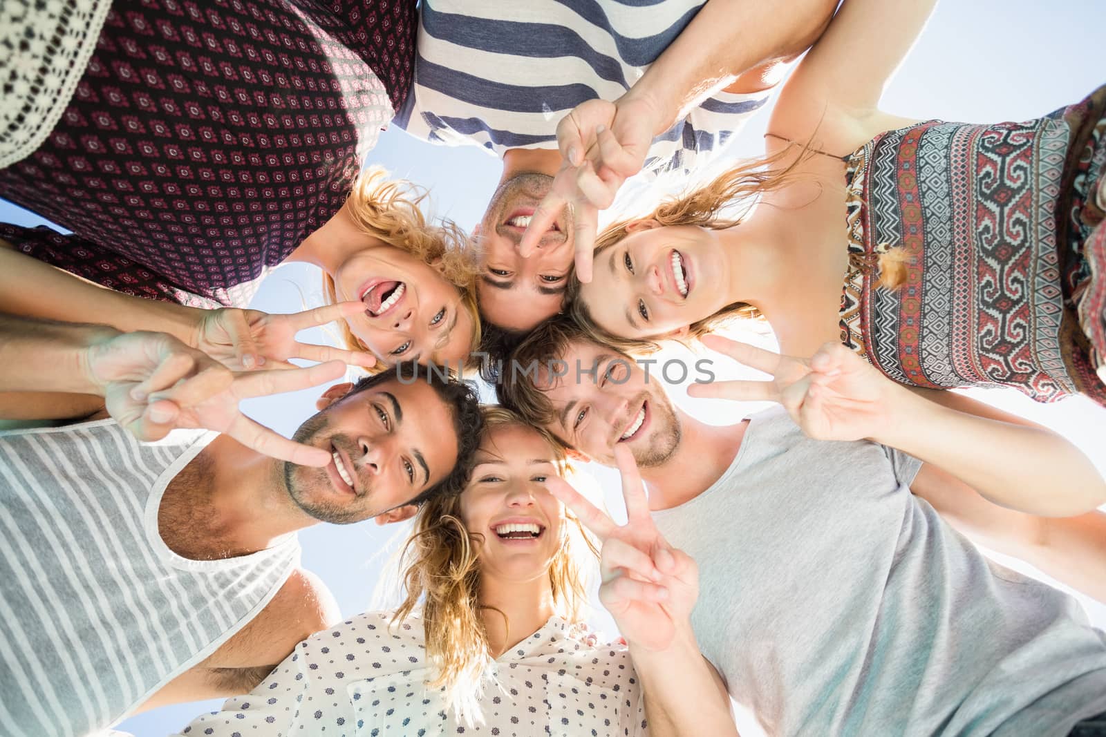 Happy friends showing v-sign finger gesture while forming a huddle against sky