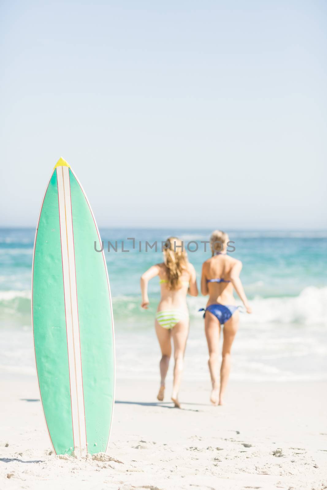 Surfboard in sand and two rear women running on the beach by Wavebreakmedia