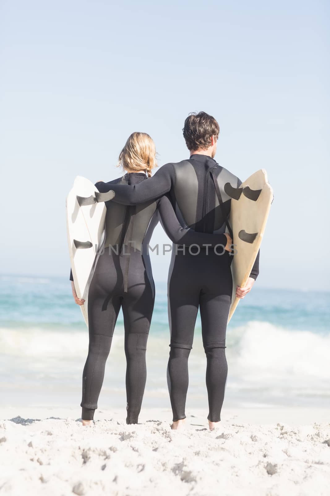 Rear view of couple with surfboard standing on the beach on a sunny day