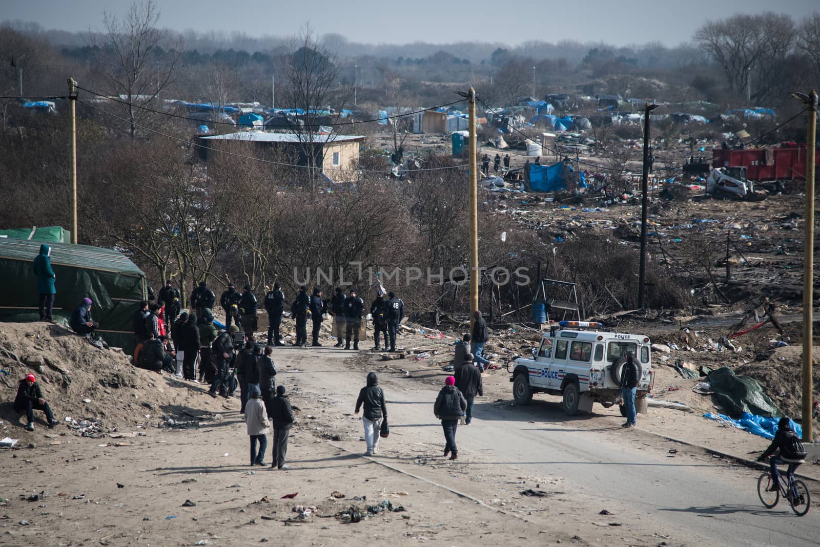 FRANCE, Calais: Migrants walk past French anti-riot policemen standing guard during the dismantling of the southern part of the so-called 'Jungle' migrant camp in Calais, northern France, on March 14, 2016.Thousands of migrants have been living in the Jungle and other smaller camps along the northern coast, desperate to reach Britain where many have family or community ties and see better hopes of gaining employment or education. 