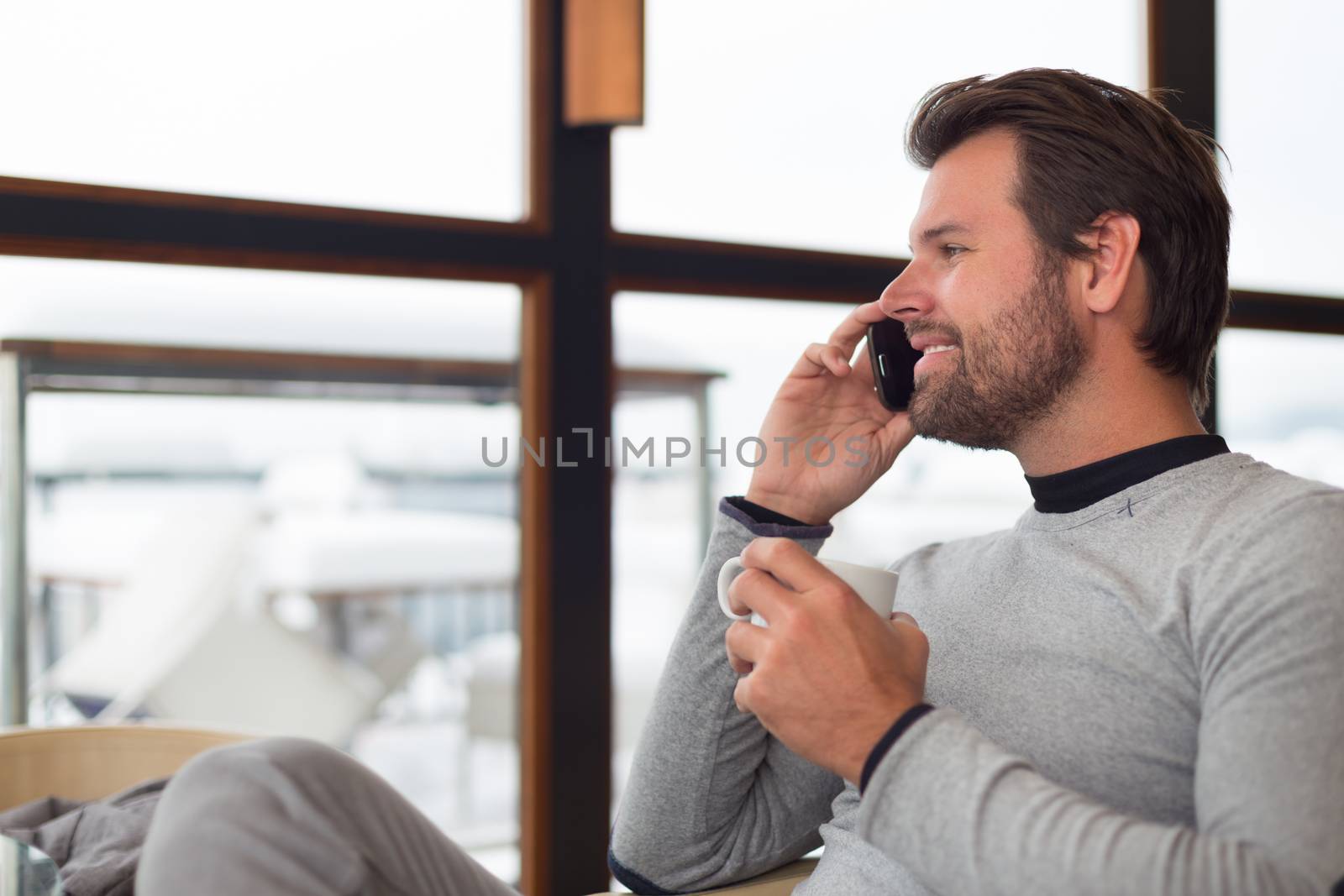 Relaxed charming man sitting in front of window with cup of morning coffee or tea near window chatting on phone.