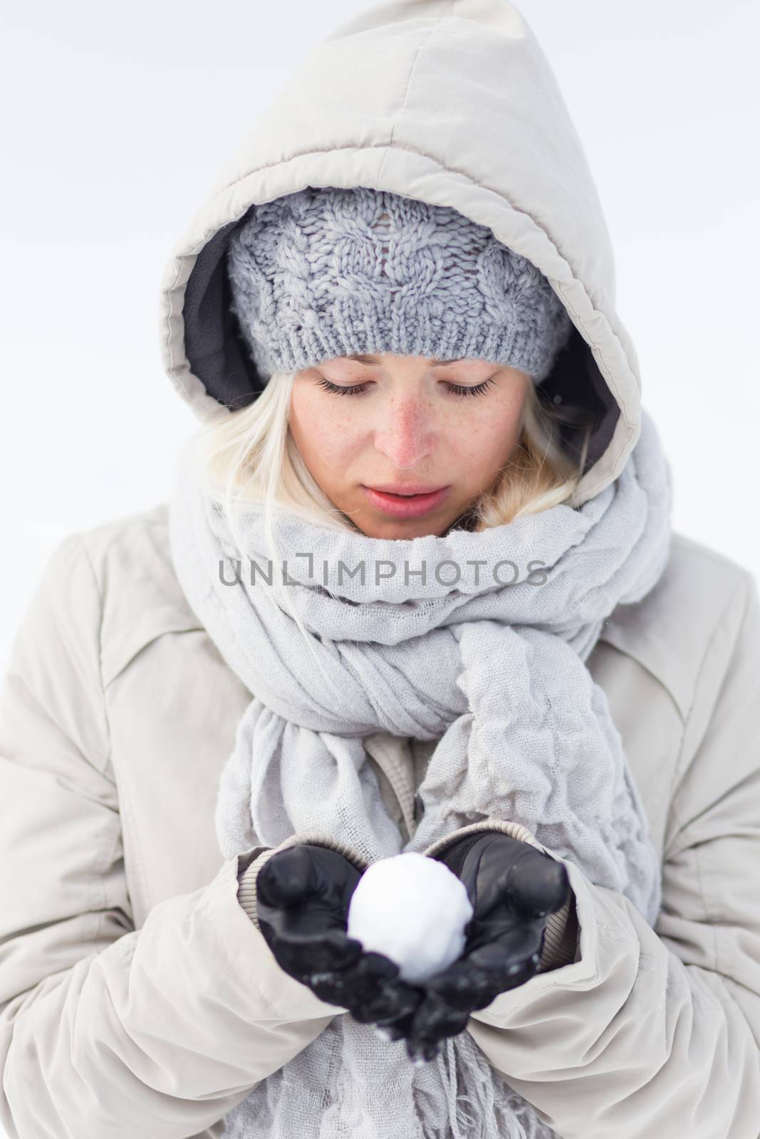 Cute casual young woman wearing glooves, woolen cap and scarf, holding icy snowball in cold winter time. Lady looking down at snowball.