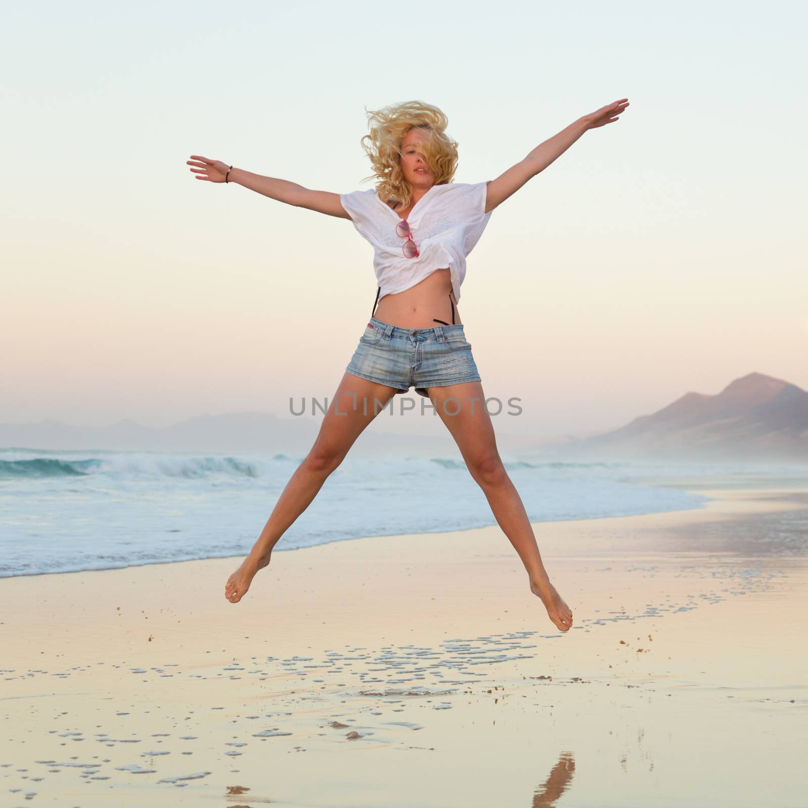 Young beautiful woman wearing jeans shorts and white t-shirt jumping on Cofete beach, Fuerteventura, Spain in sunset.