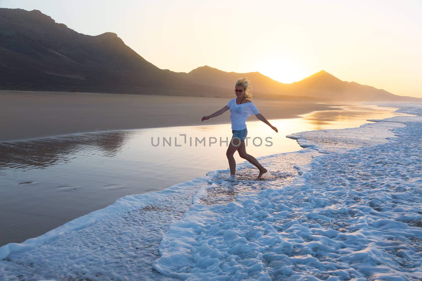 Woman having fun runing from waves on solitary sandy beach in sunset. Waves sweeping away her traces in sand. Beach, travel, concept. Copy space. 