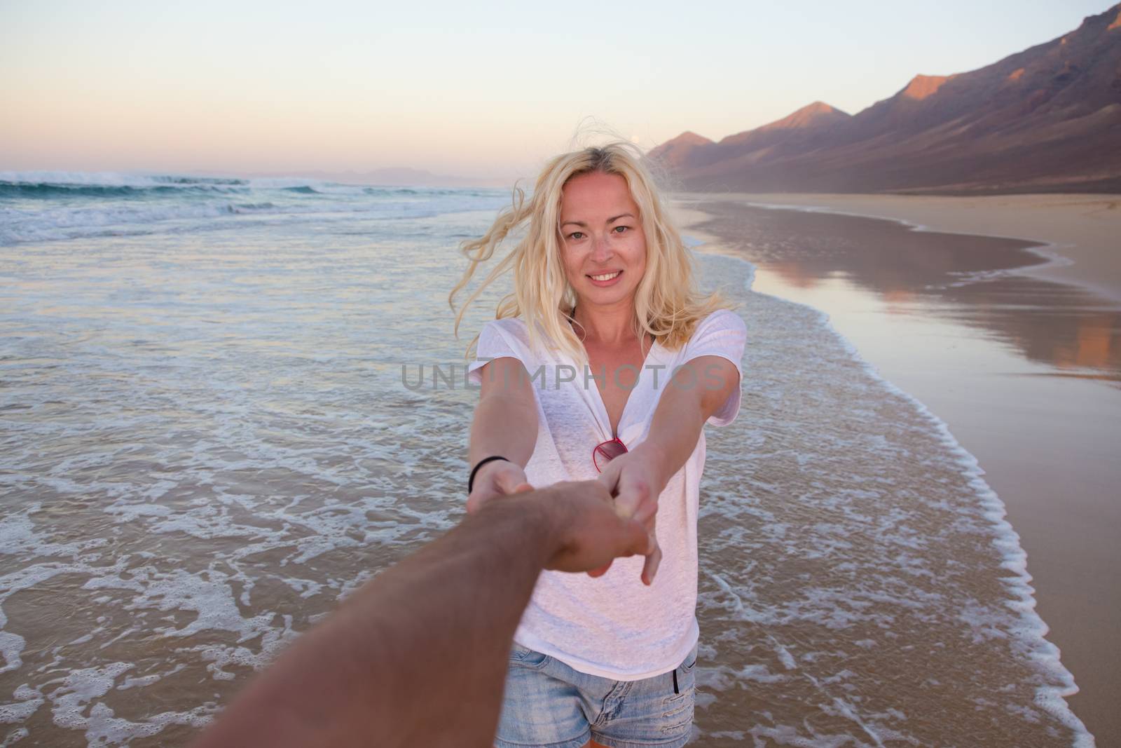 Young romantic couple, holding hands, having fun on perfect deserted beach at sunset. Shot from boyfrieds perspective. Guy looking at her beautiful carefree girlfriend.