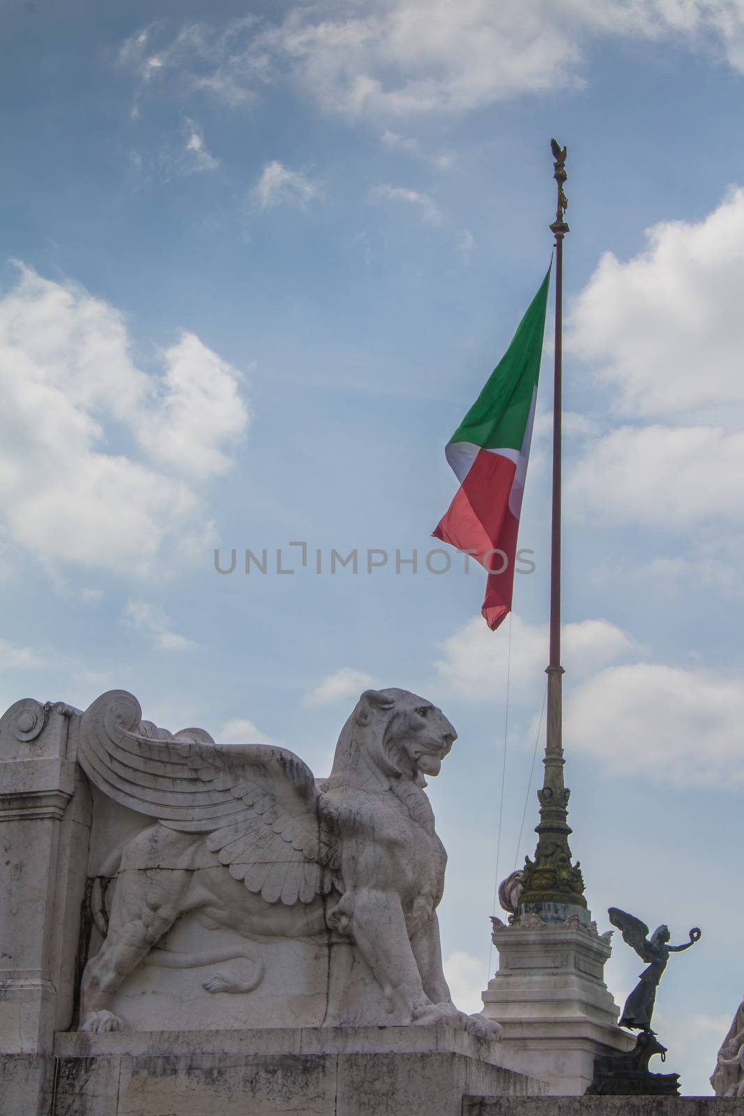 Detail of the Vittorio Emanuele Monument in Italy, Rome, Piazza Venezia. Statue of a lion with wings with an italian flag in the background. Cloudy sky.