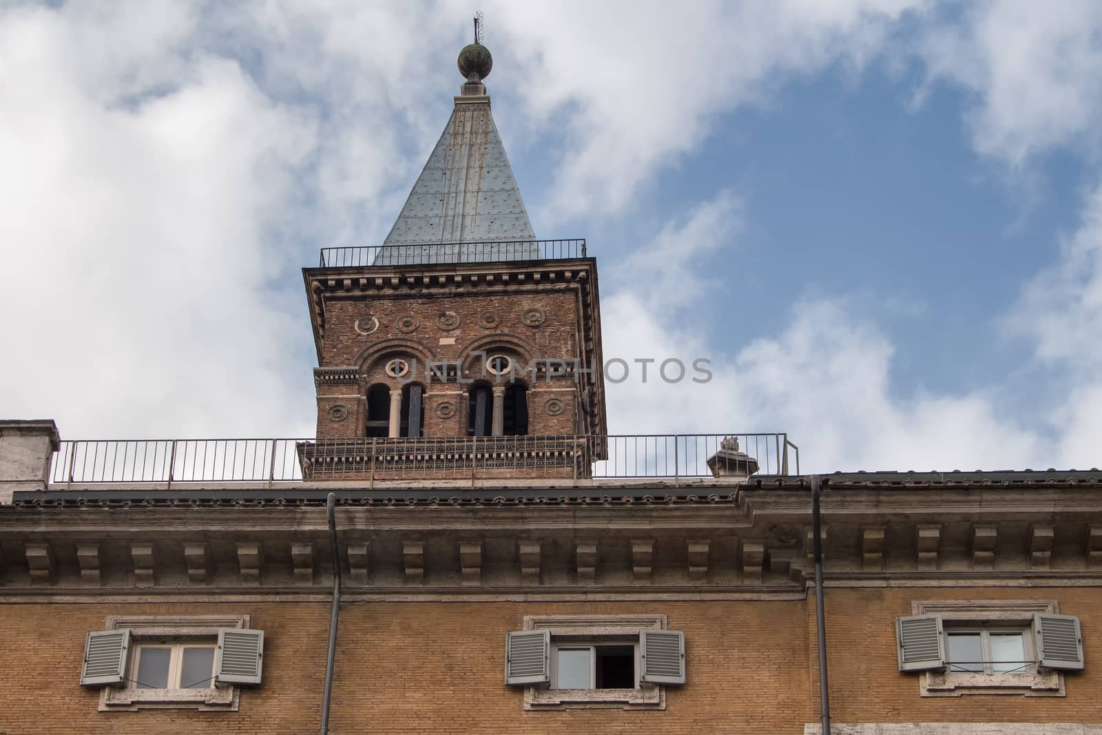 Typical orange colored building with small windows with shutters in Rome. Behind a tower of a church. Cloudy sky.