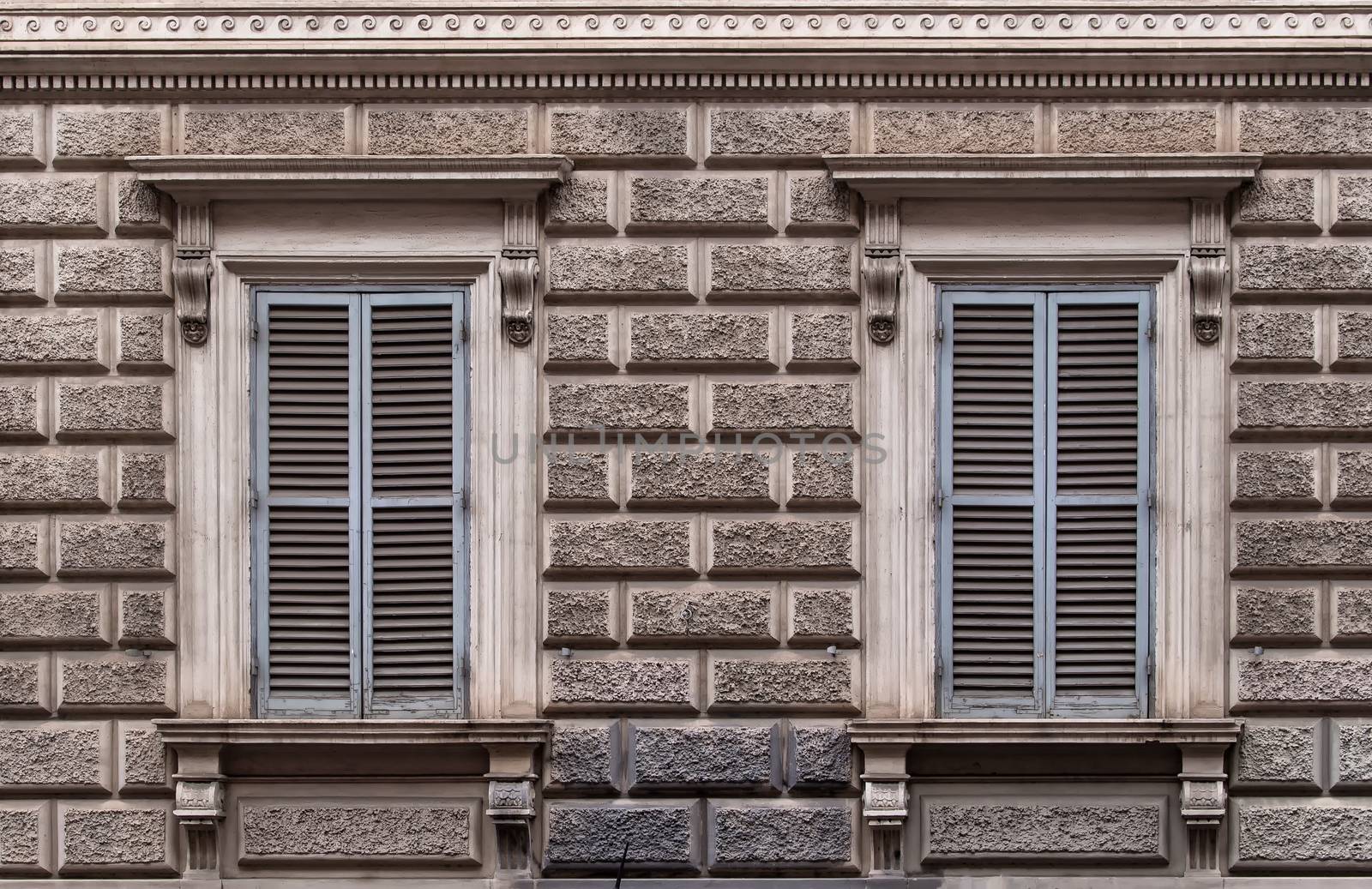 Facade of the building decorated to a design of big stone bricks. Two windows with grey closed shutters. Rome, Italy.