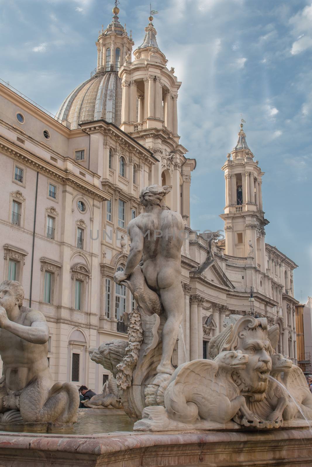 View from the back part on the statue at the fountain and Church Sant' Agnese in Agone. Cloudy sky with rays of sun.