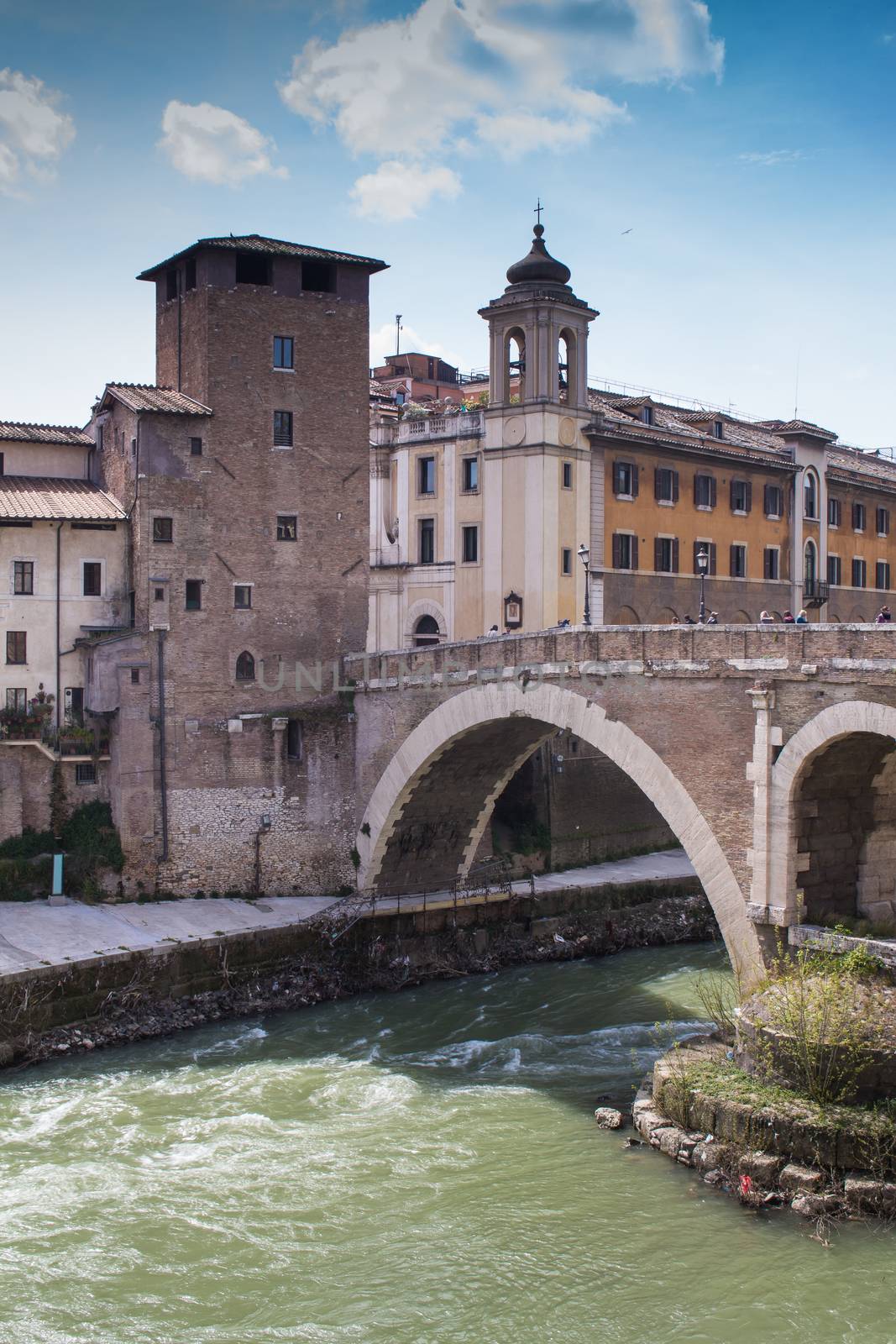 Green water of river Tiber, surrounding the Island with historical buildings and an old bridge. Cloudy blue sky.