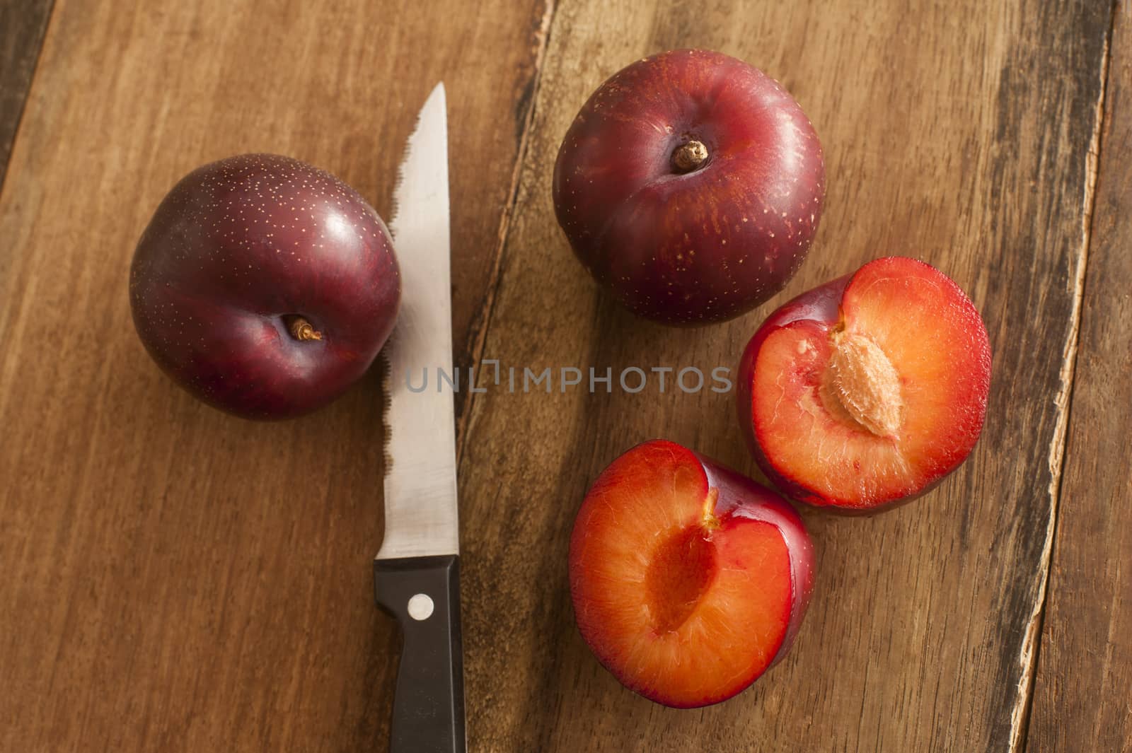 Fresh whole and halved plums showing the stone or pip lying on a wooden table with a knife in a close up overhead view