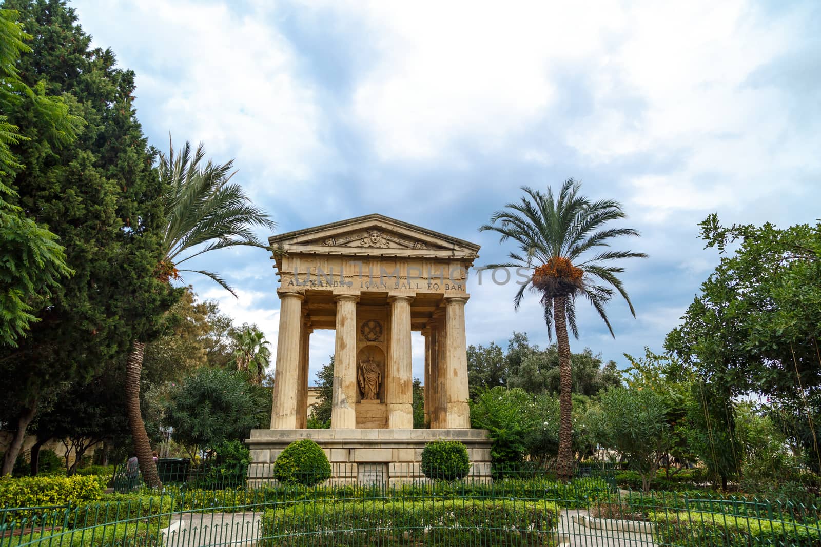 View of Lower Barrakka Gardens with historical monumental columns in Malta, with small trees around, on cloudy blue sky background.