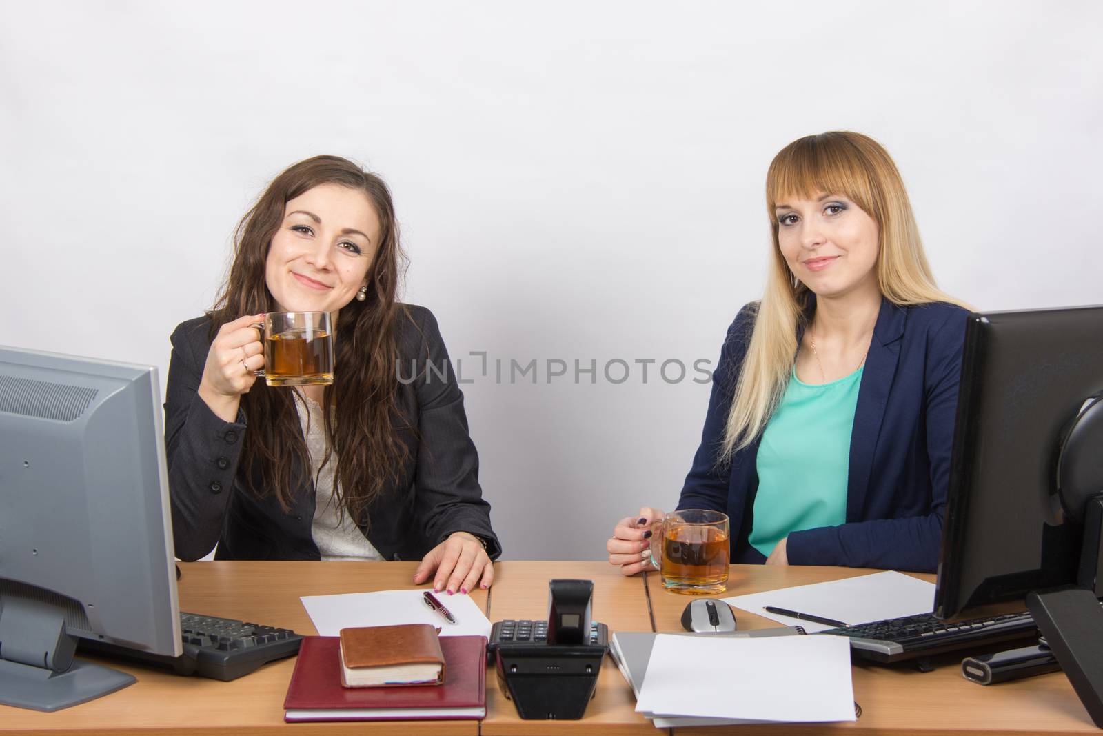 Two young beautiful girls drink tea, office behind a desk in transparent glasses