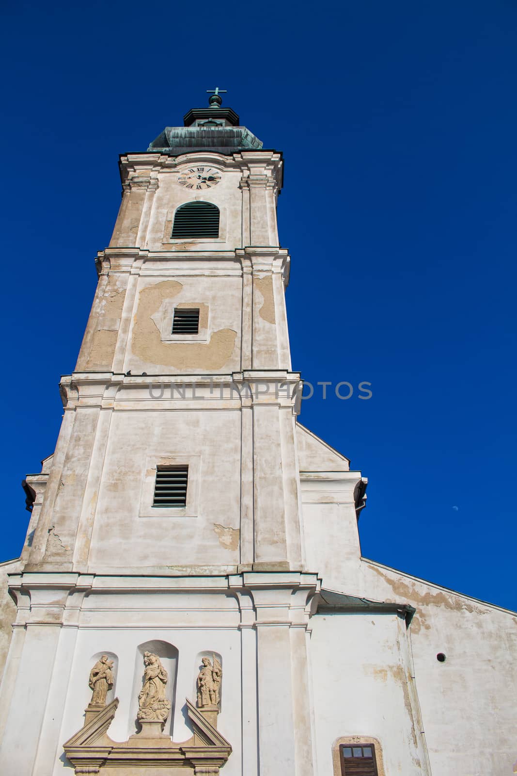 Church of Saint Cross. Old facade, high tower with clock. Cross on the tower. Bright blue sky.