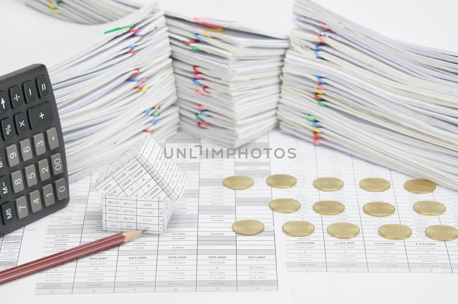 House and brown pencil on finance account have row of gold coins and calculator place vertical with overload of paperwork with colorful paperclip as background.