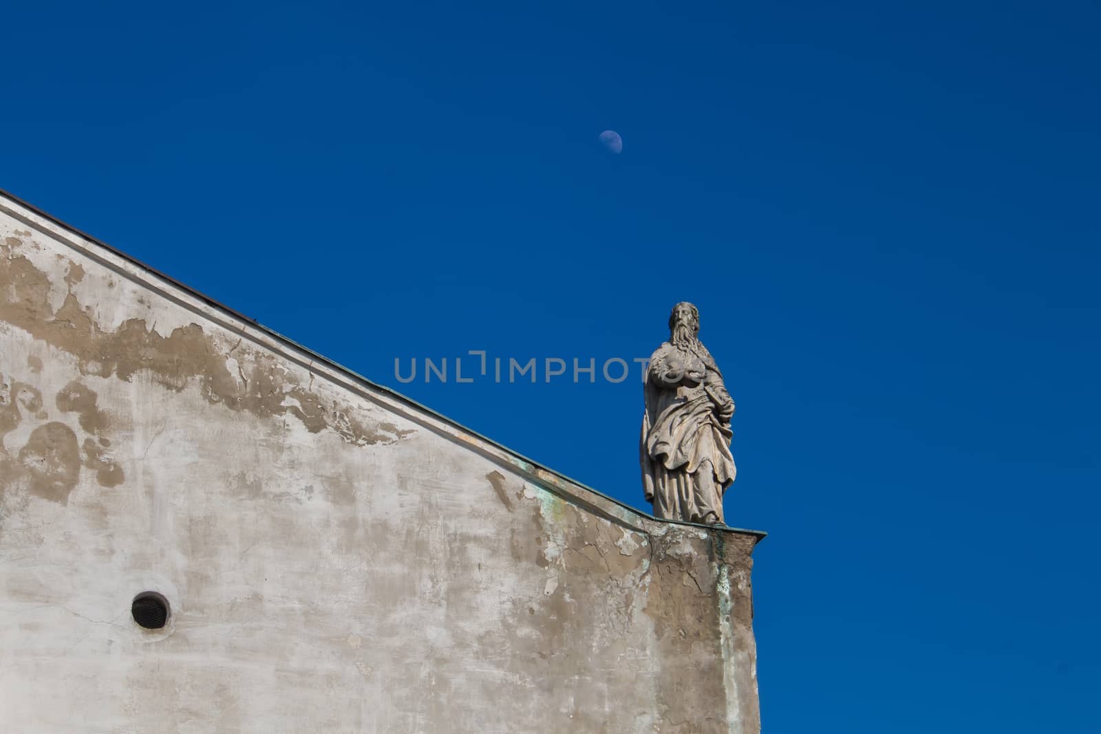 Diagonal line of the roof of the Church of Saint Cross in Devin, Bratislava, Slovakia. Statue of a saint. Bright blue sky with a moon.