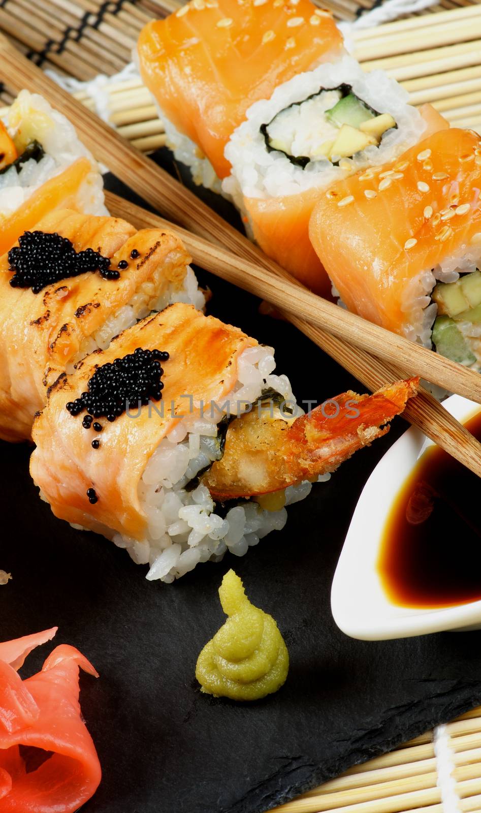 Arrangement of Maki Sushi with Smoked Salmon and Eel with Black Caviar on Stone Plate with Soy Sauce, Ginger and Pair of Chopsticks closeup on Straw Mat background