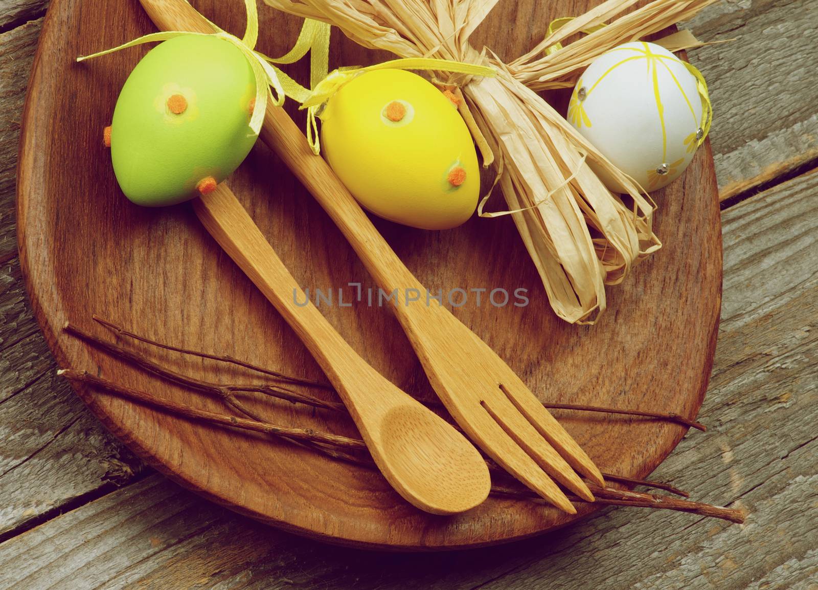 Easter Table Setting with Wooden Fork and Spoon, Colored Eggs and Natural Branches on Wooden Plate closeup on Rustic Wooden background. Retro Styled