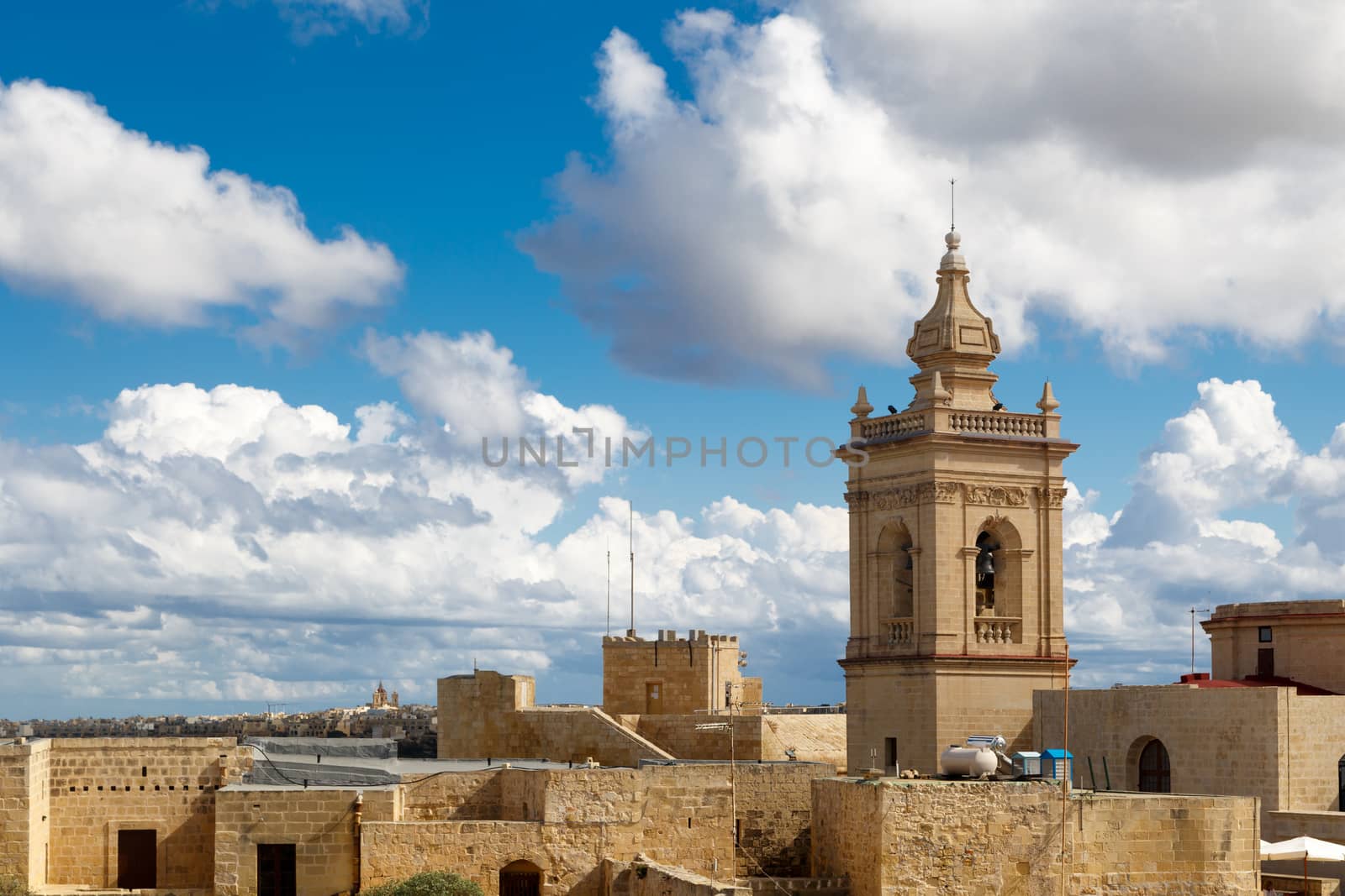 View of a historical church in Victoria, Malta with a limestone belltower, on cloudy sky background.