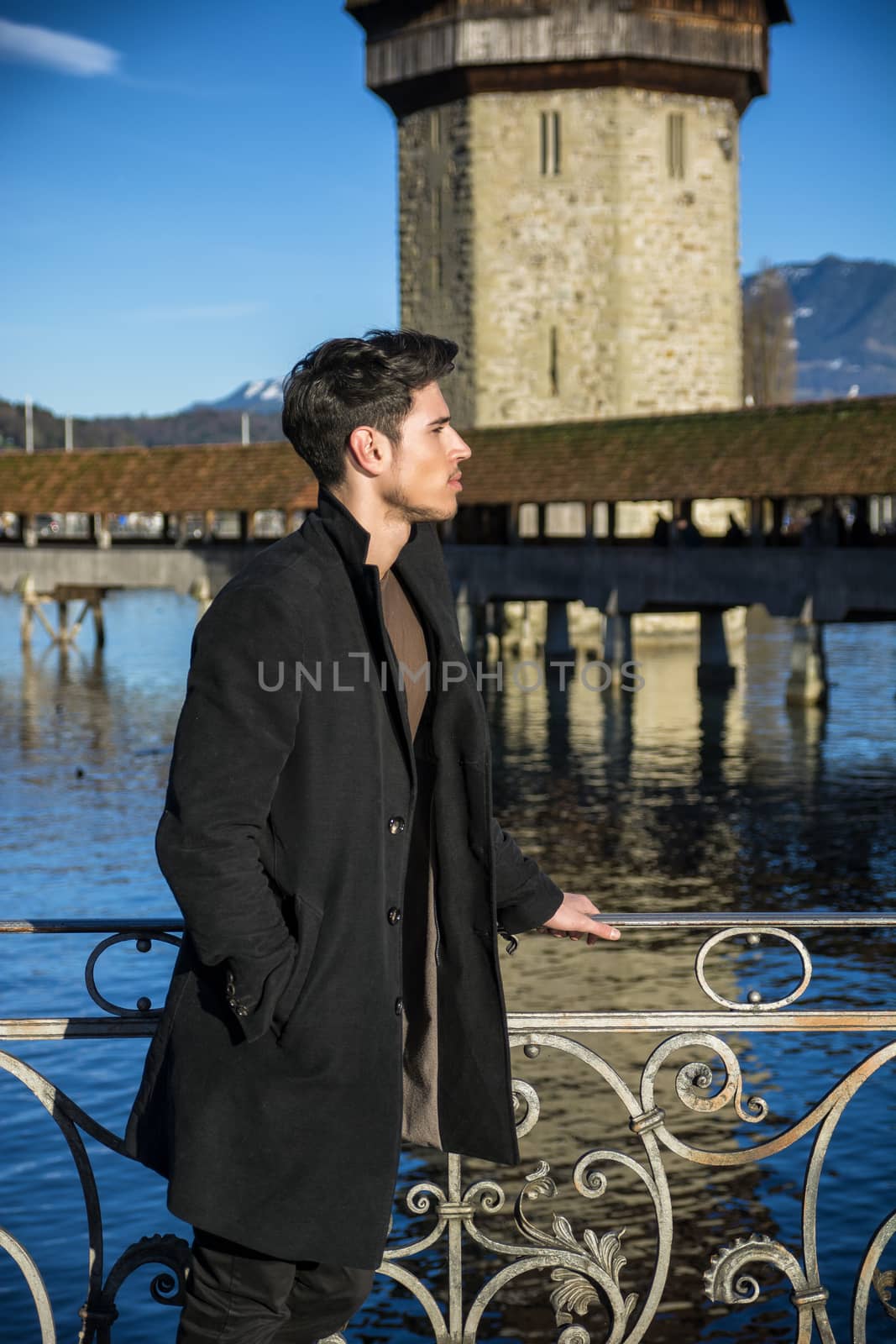 Handsome man standing near metal fence in Lausanne. Famous wooden bridge and Water Tower on background.