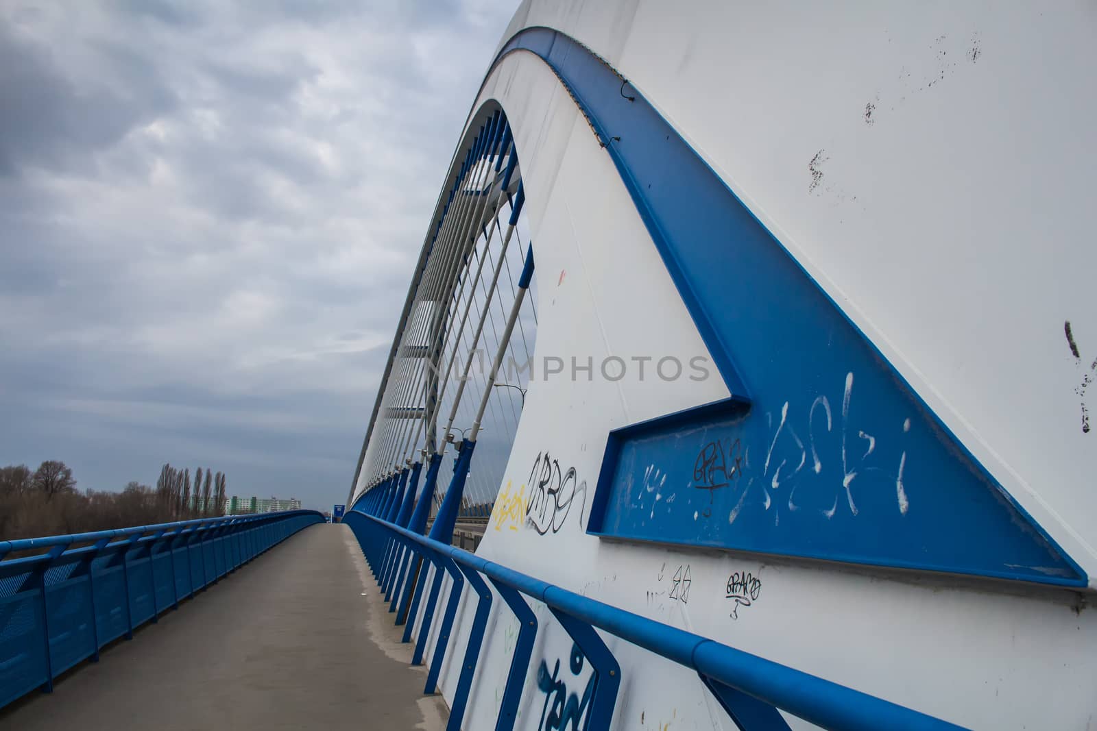 One of the bridges in slovak capital Bratislava. Grey and blue lines and arch of the modern Apollo bridge and a patway. Cloudy sky.