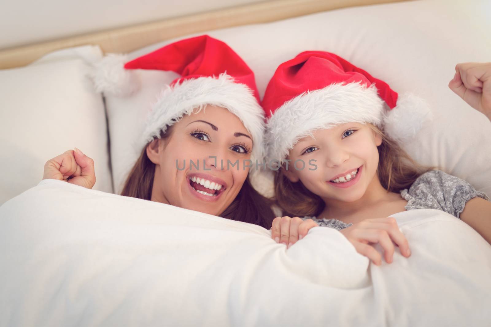 Beautiful smiling mother and her daughter with santa hat having fun in bed. They lie in bed looking at camera and peeking under the blanket with smile.