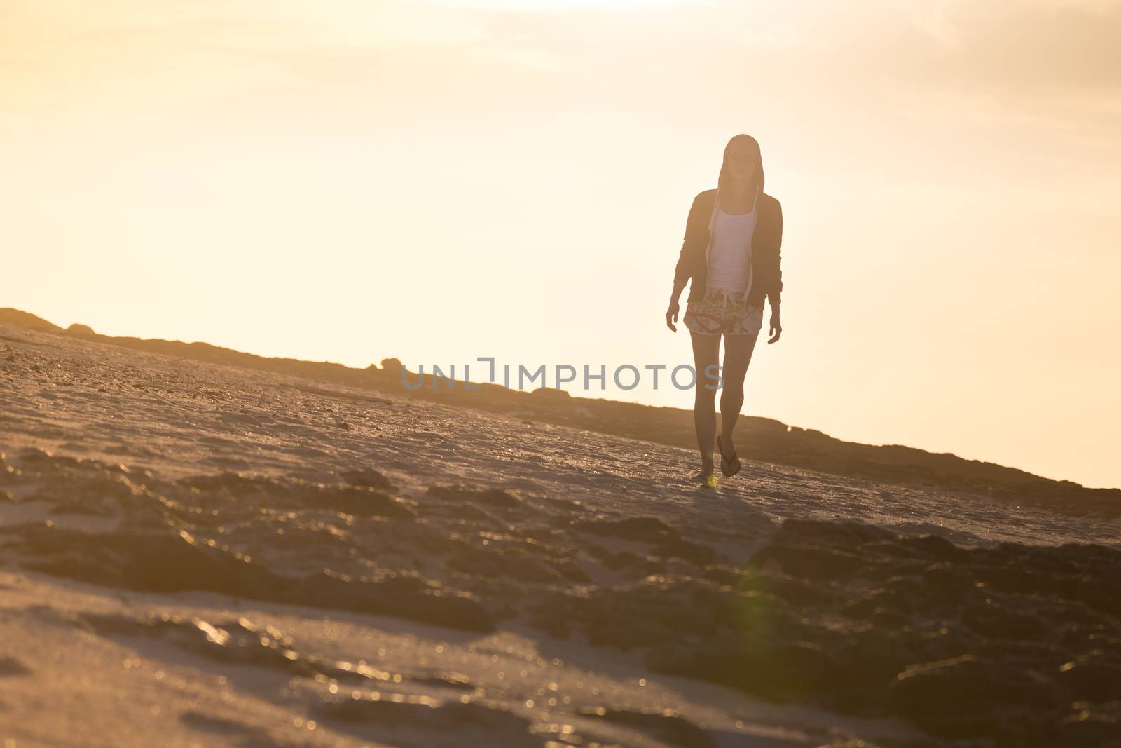 Casualy dressed active sporty blonde woman walking on rocky beach in sunset.