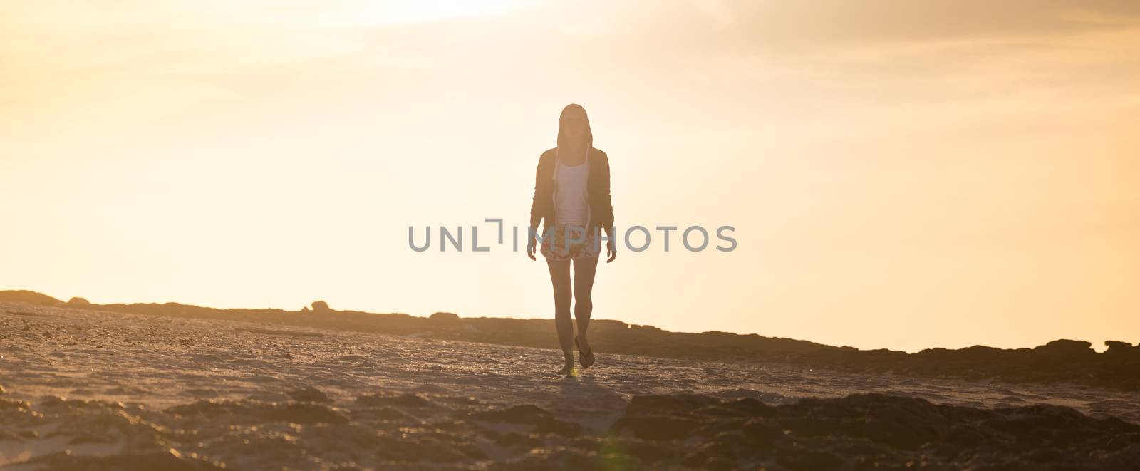 Casualy dressed active sporty blonde woman walking on rocky beach in sunset.