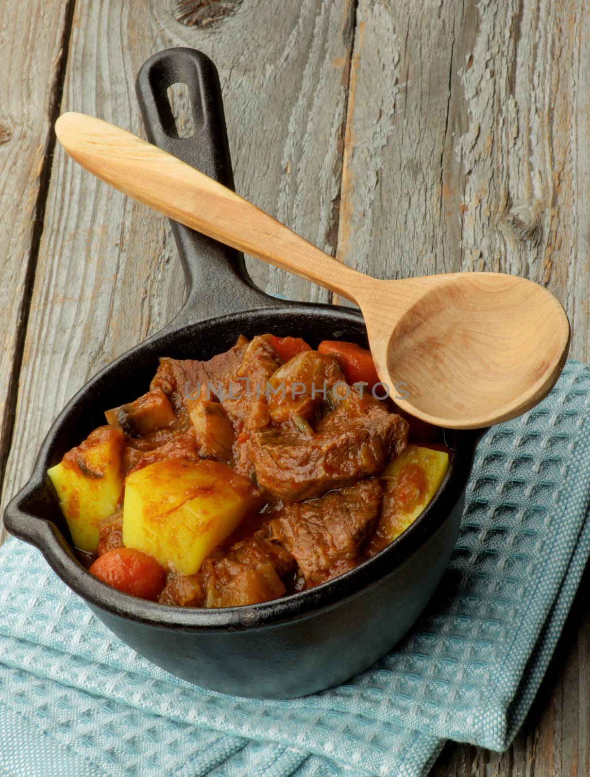 Homemade Beef Stew with Carrots, Potatoes, Celery and Leek in Cast Iron on Blue Napkin with Wooden Spoon closeup on Wooden background