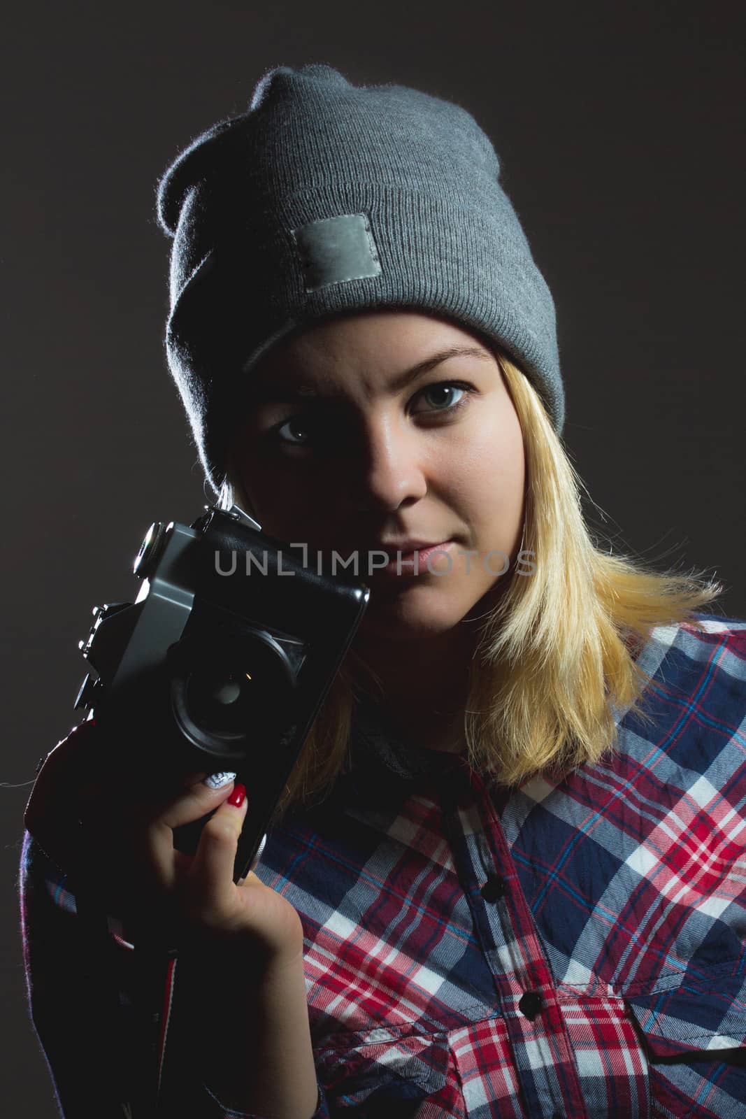 Portrait of hipster girl posing with retro camera wearing checkered shirt and winter hat on dark background