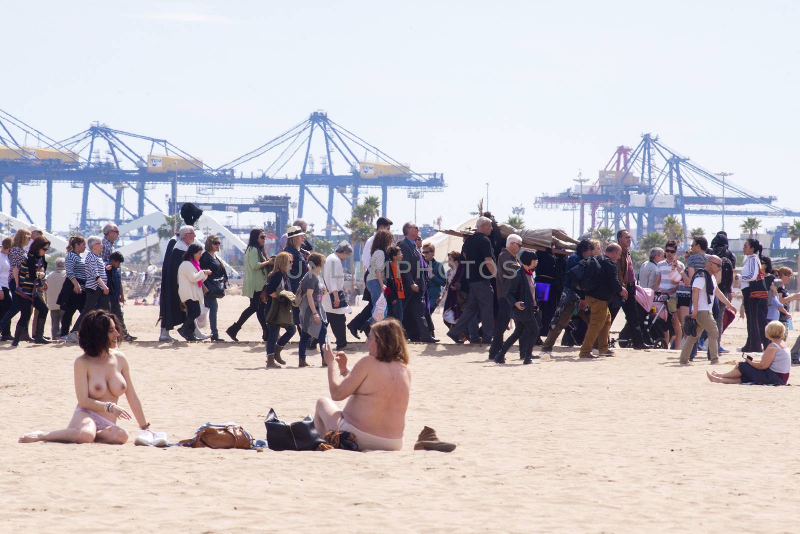 SPAIN, Valencia: Women sunbathe as penitents of the “Cristo Salvador y del Amparo” (Christ the Saviour and Protector) brotherhood take part in the Good Friday procession on Malvarrosa beach, in Valencia, on March 25, 2016. Christian believers around the world mark the Holy Week of Easter in celebration of the crucifixion and resurrection of Jesus Christ.