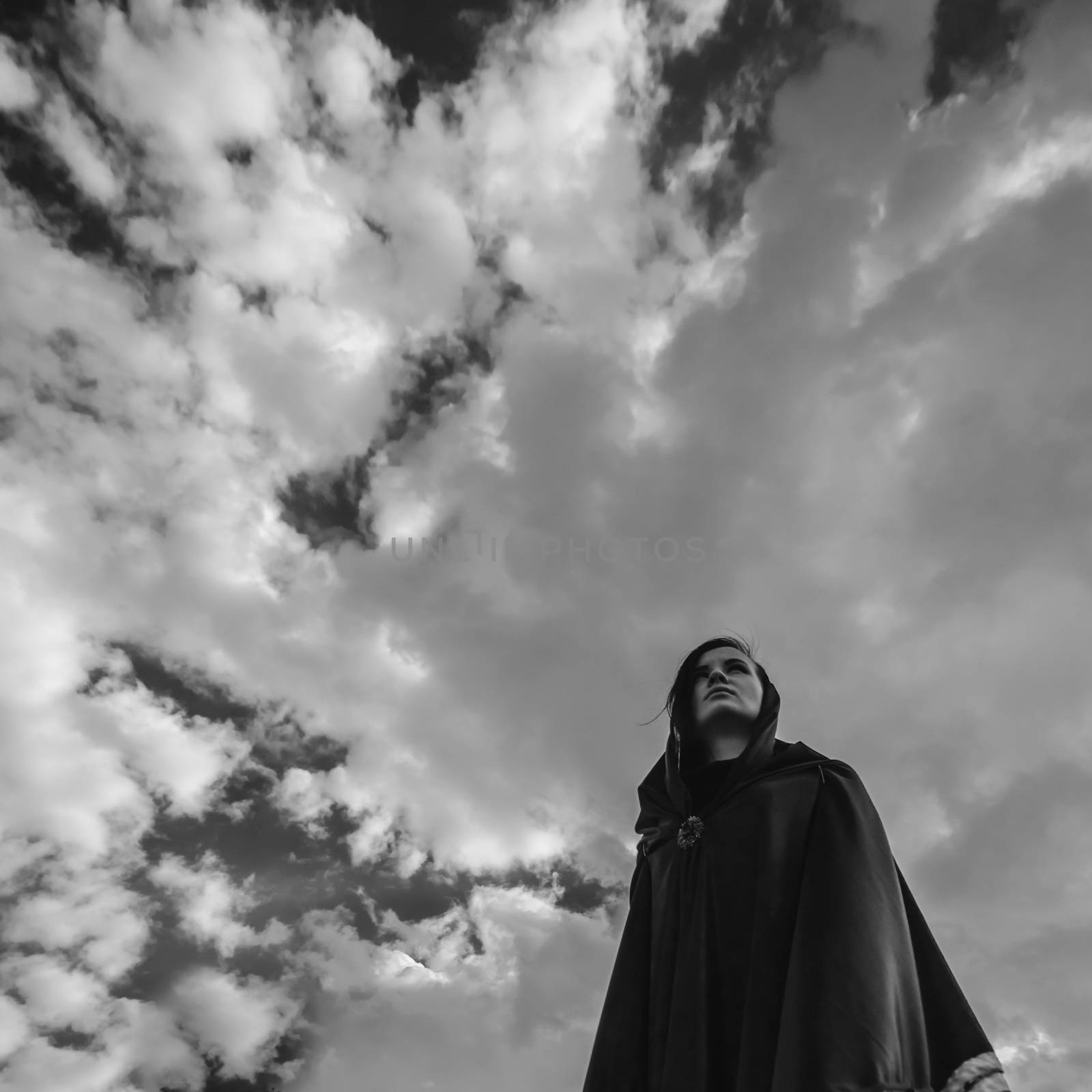 a young witch in a black robe with a hood on the background of dramatic sky
