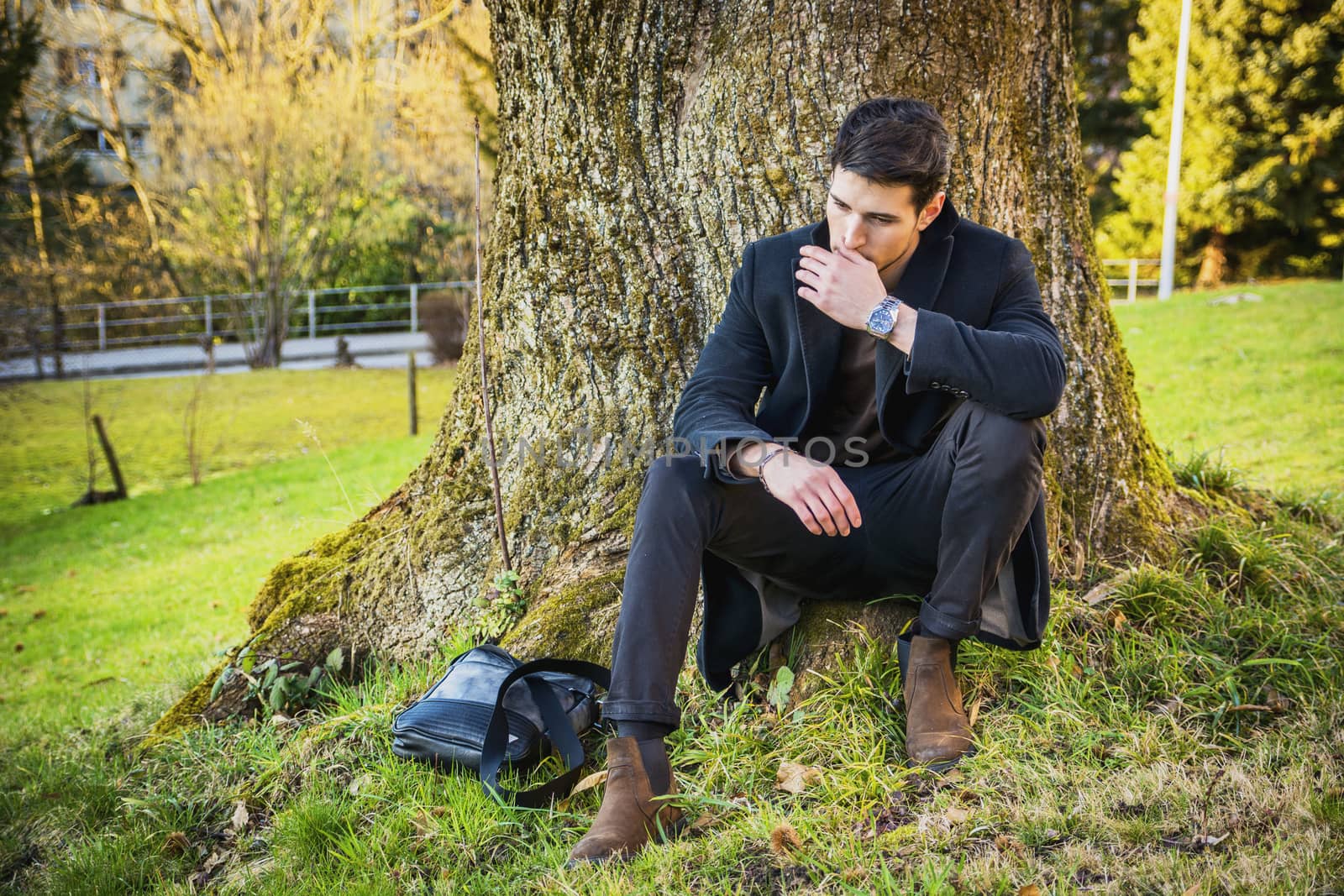 Handsome young man leaning against tree, looking to a side, in a sunny day wearing a black coat