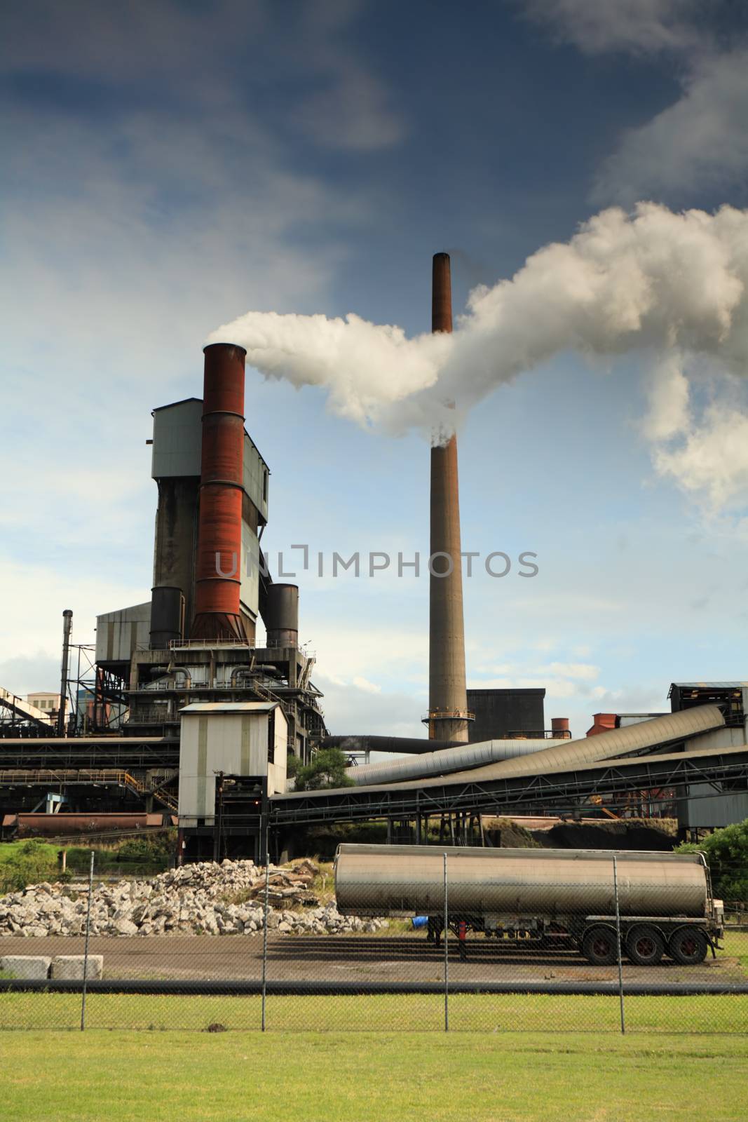 Steel mill smelter emitting toxic fumes and air pollutants billowing up and out of one of its many tall chimneys.  Pollutants can vary and include hydrogen fluoride, sulfur dioxide, oxides of nitrogen, offensive and noxious smoke fumes, vapors, gases, and other toxins. A variety of heavy metals: lead, arsenic, chromium, cadmium, nickel, copper, and zinc are also released by the facilities.